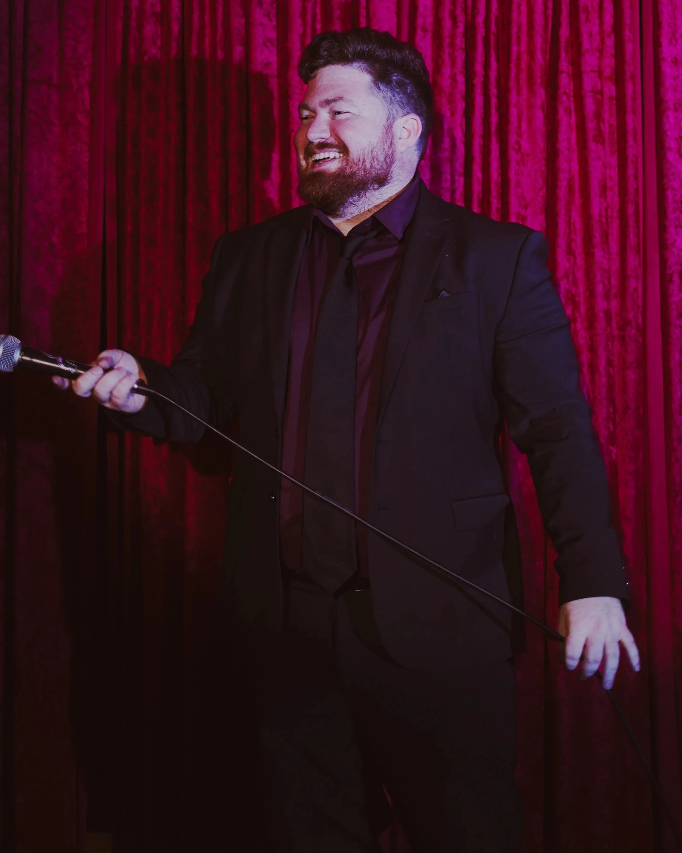 A man in a black suit holding a microphone, smiling, standing in front of a red velvet curtain.