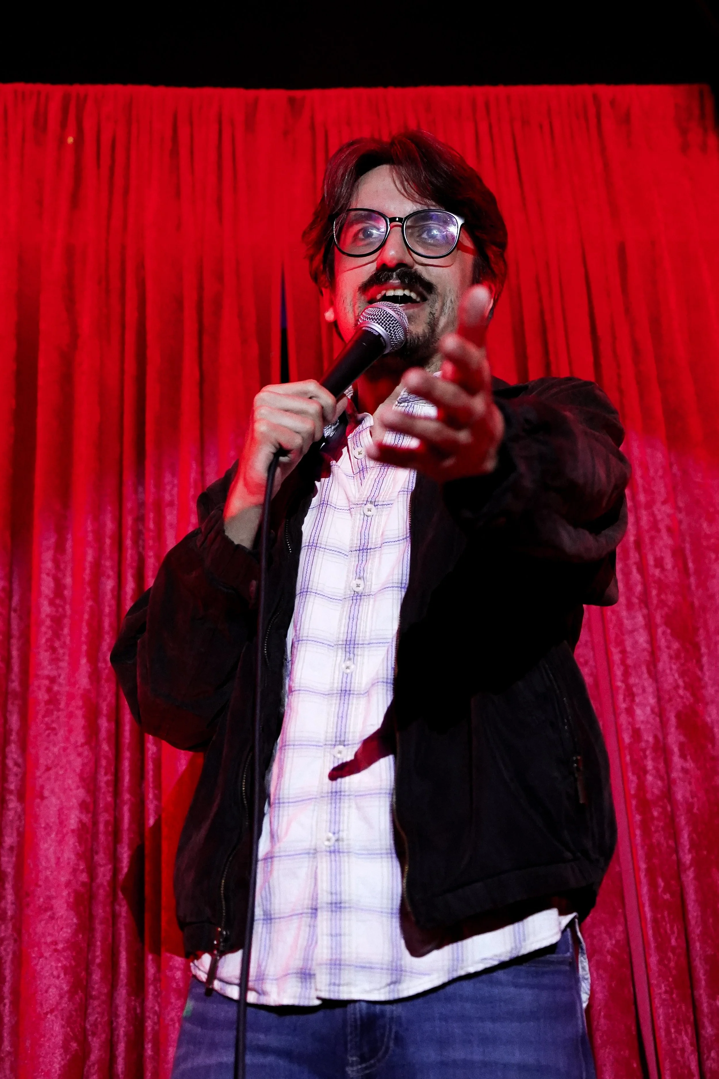 A man with glasses and a beard speaking into a microphone on a stage with red curtains in the background.