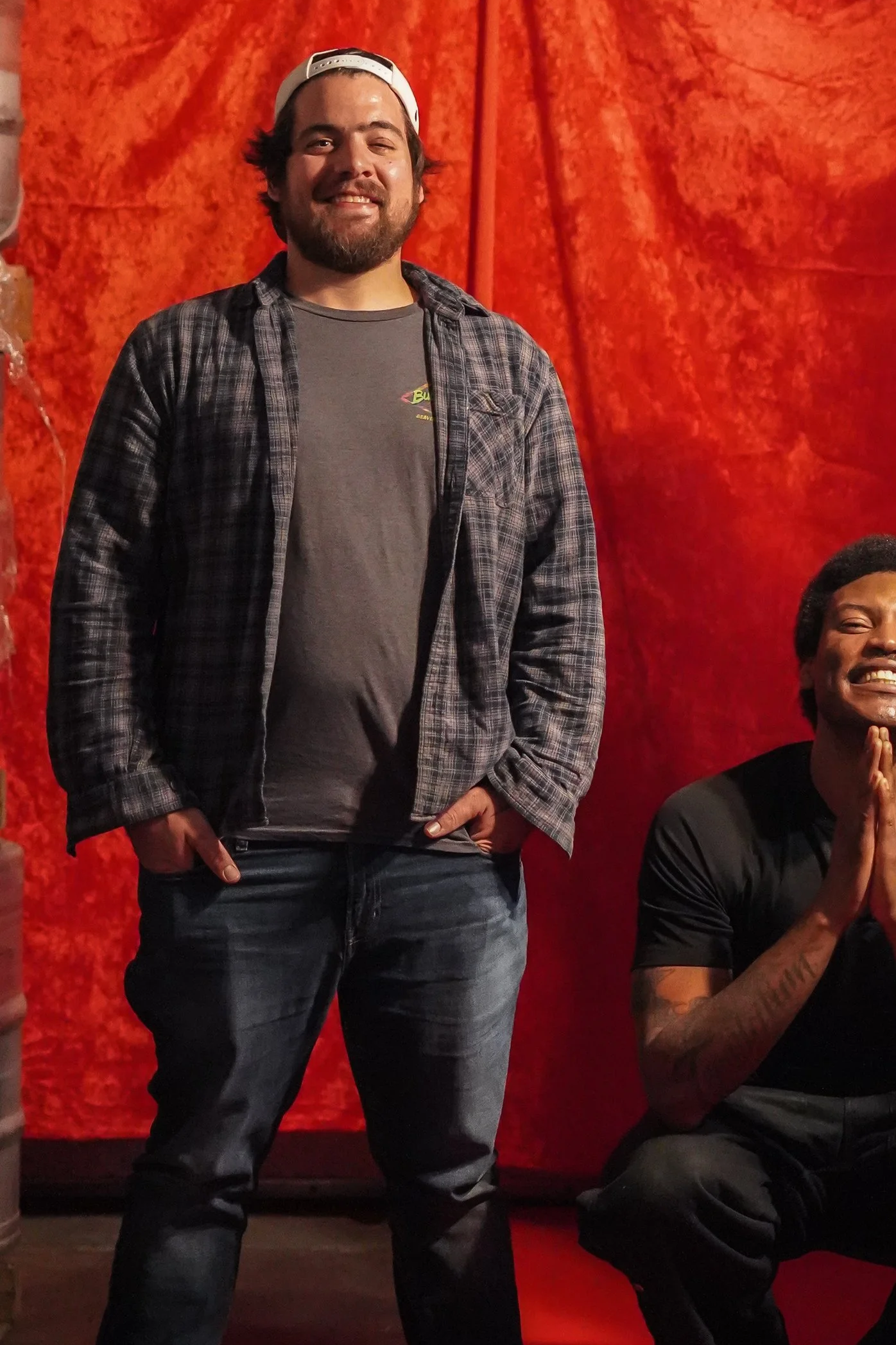 A man with a beard wearing a backwards white baseball cap, a gray T-shirt, and a checkered flannel shirt standing in front of a red textured curtain. He is smiling with his eyes partly closed.