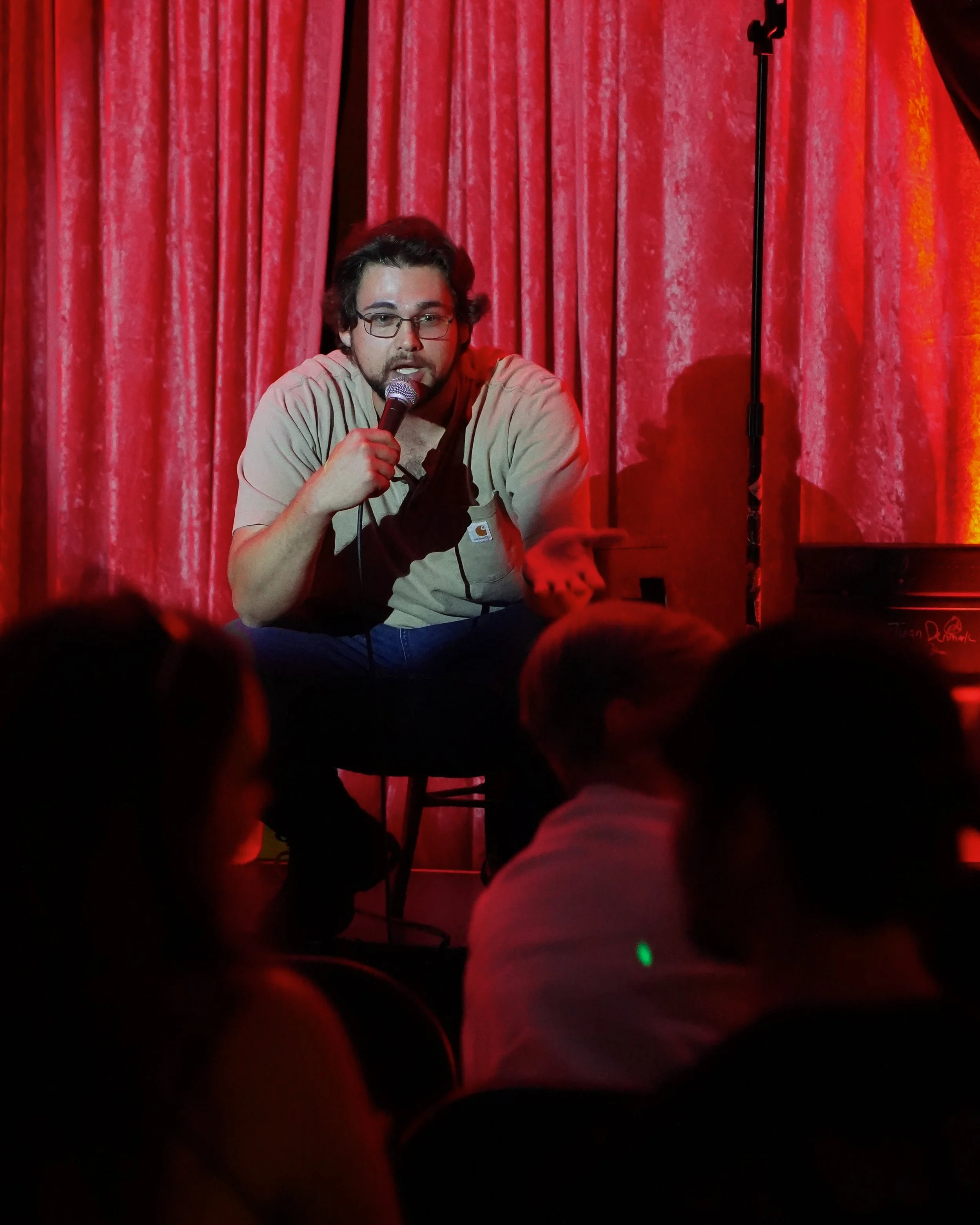A man with glasses and a beard speaking into a microphone on stage with red curtains in the background, audience in the foreground.