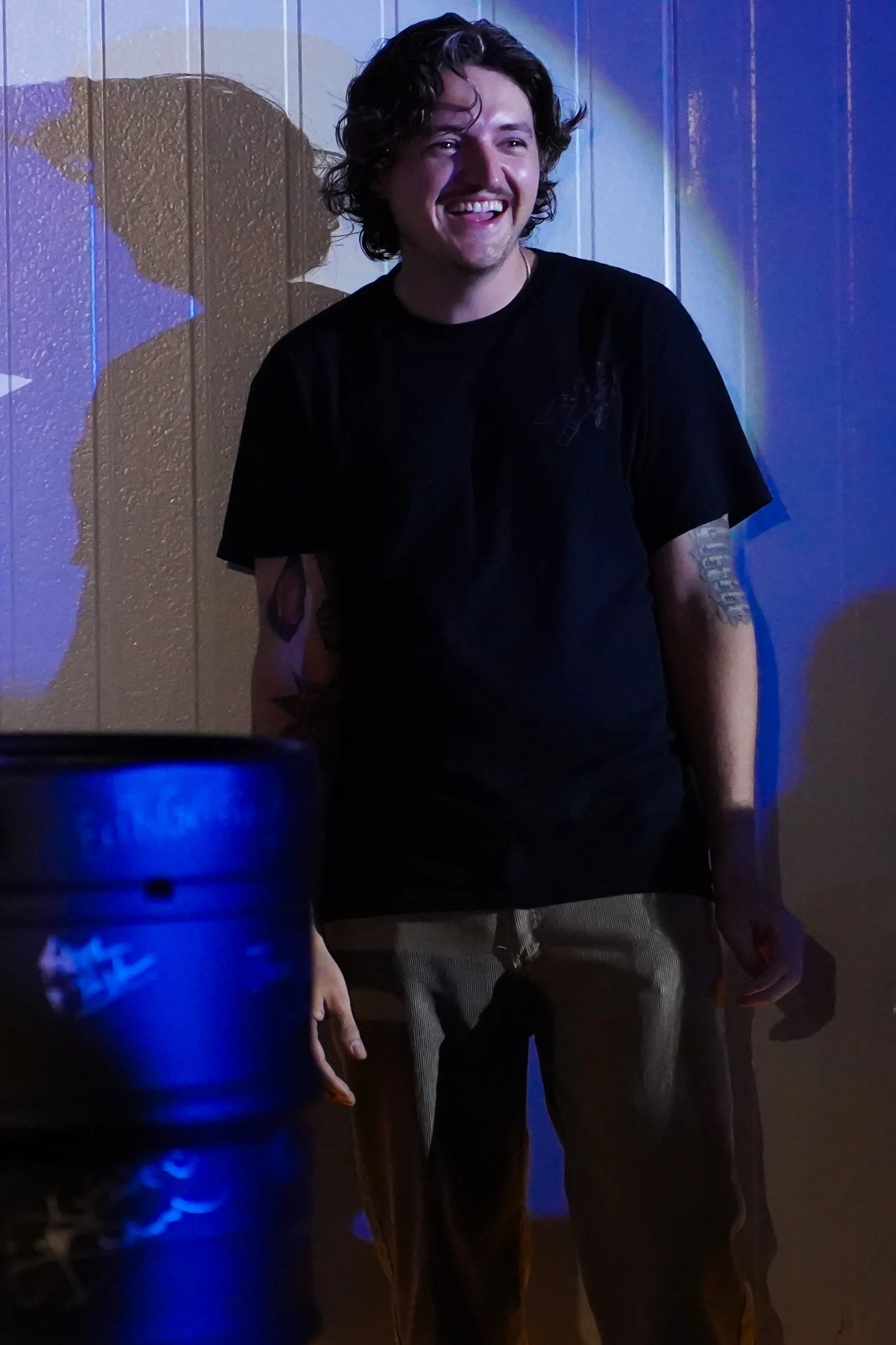 A man with curly hair smiling in front of a wooden wall, with colored lighting casting shadows on the wall.