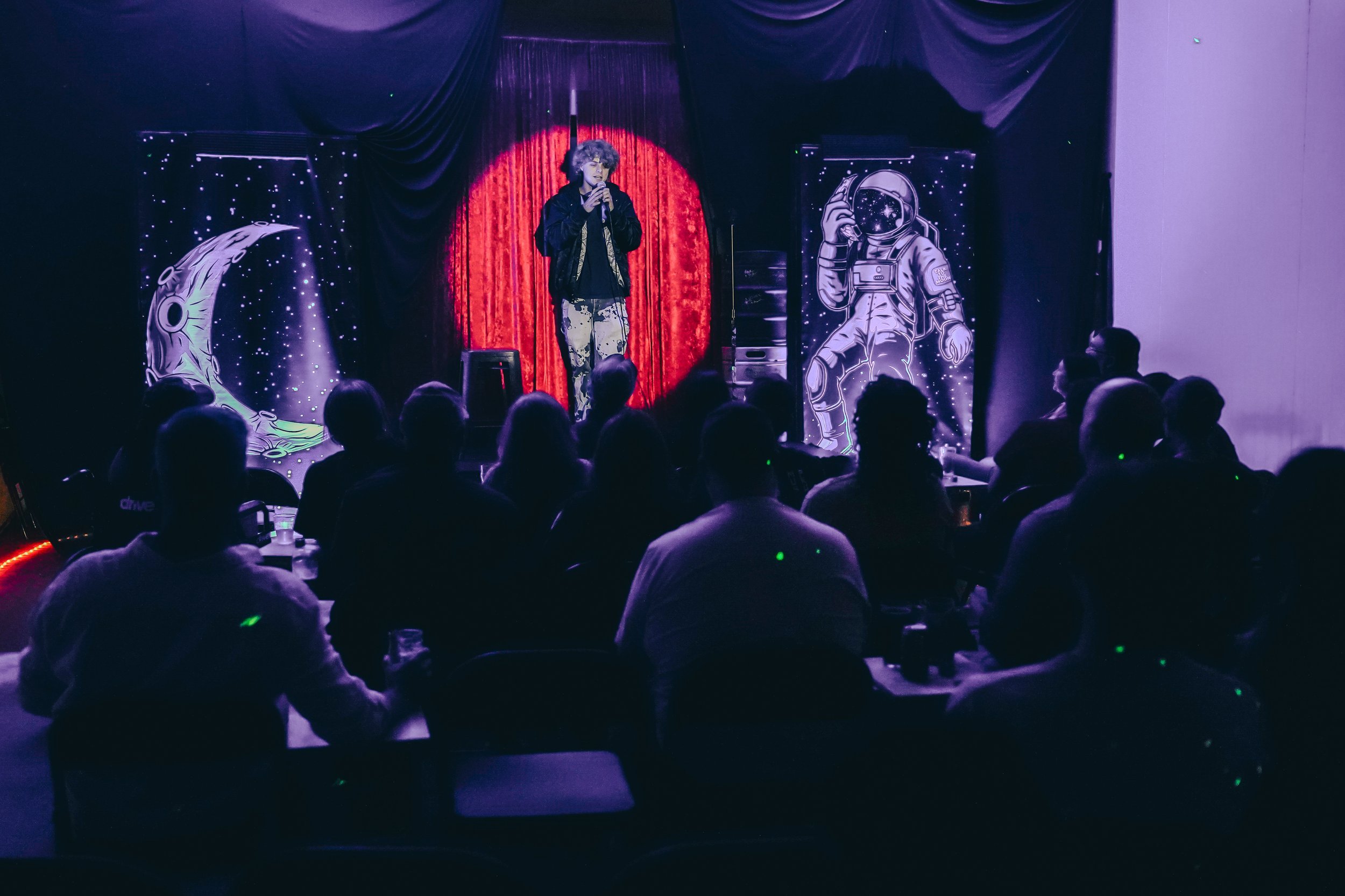 A performer on stage holding a microphone, with a red curtain and space-themed illustrations of an astronaut, moon, and stars behind them. Audience members are seated and watching the performance.