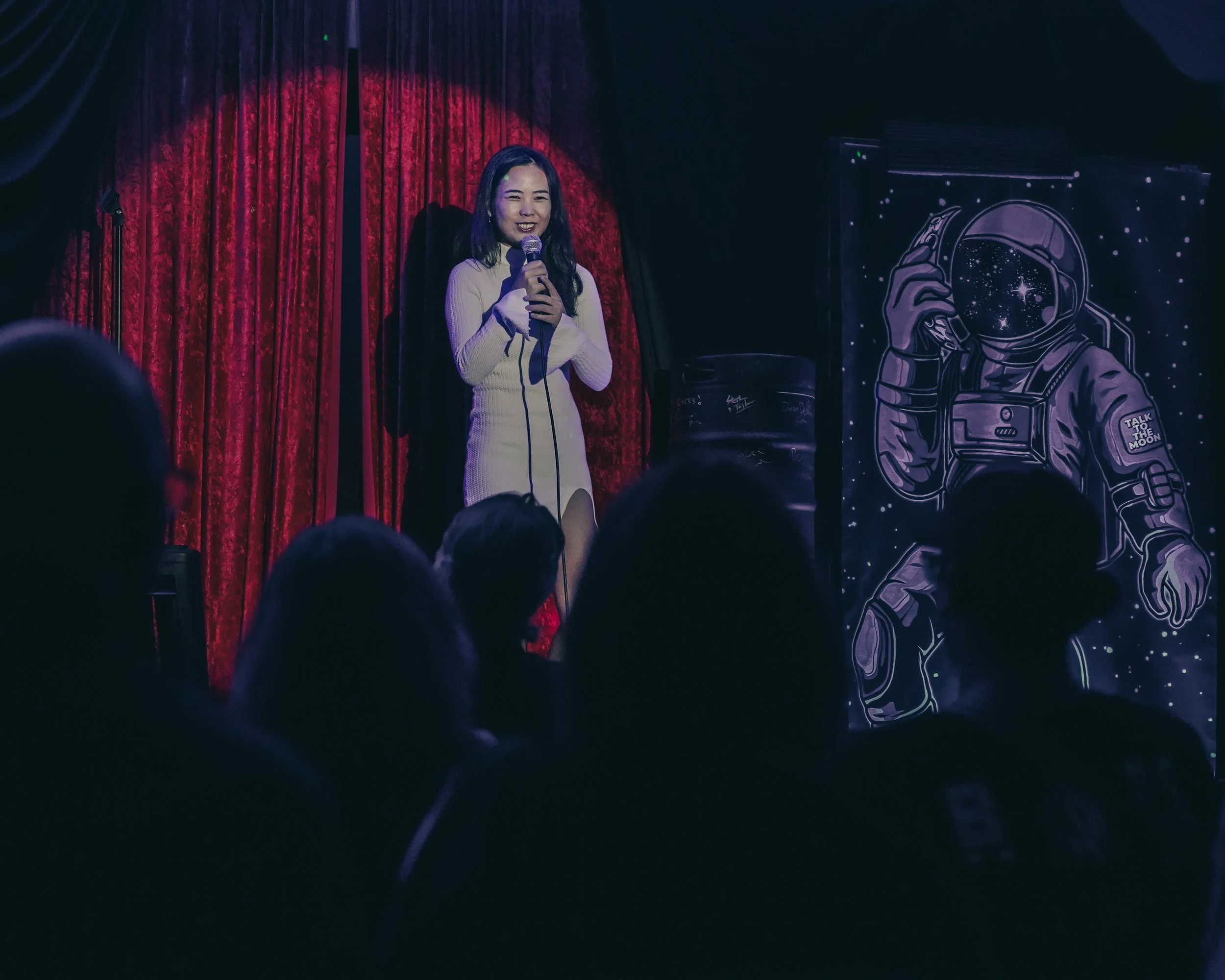 A woman dressed in white on stage holding a microphone, smiling, with red curtains behind her and space-themed artwork to the right.