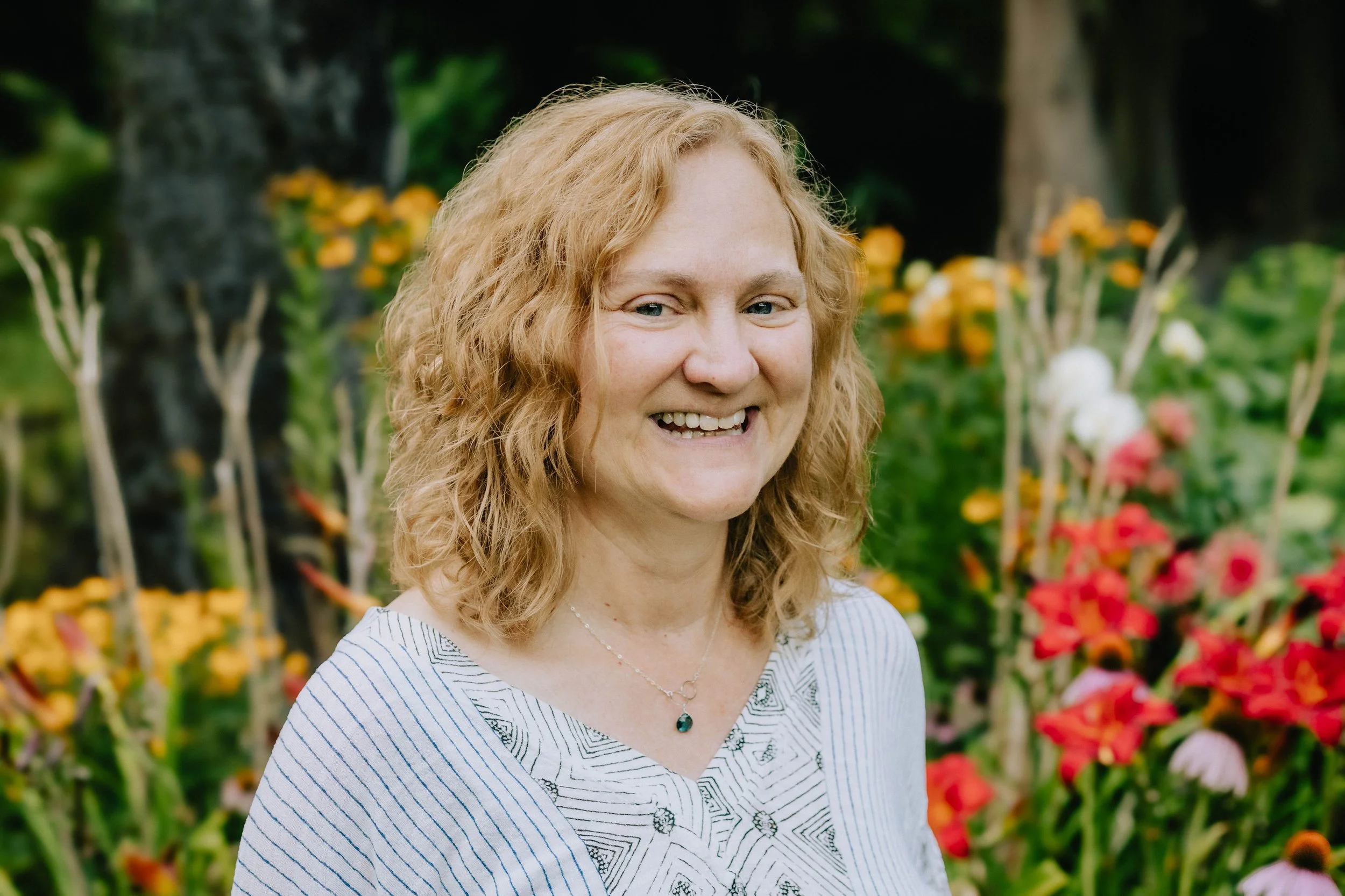 Photo of Jaye Matthews, a curly haired white woman smiling while looking into the camera in front of a blurred background of yellow and red flowers