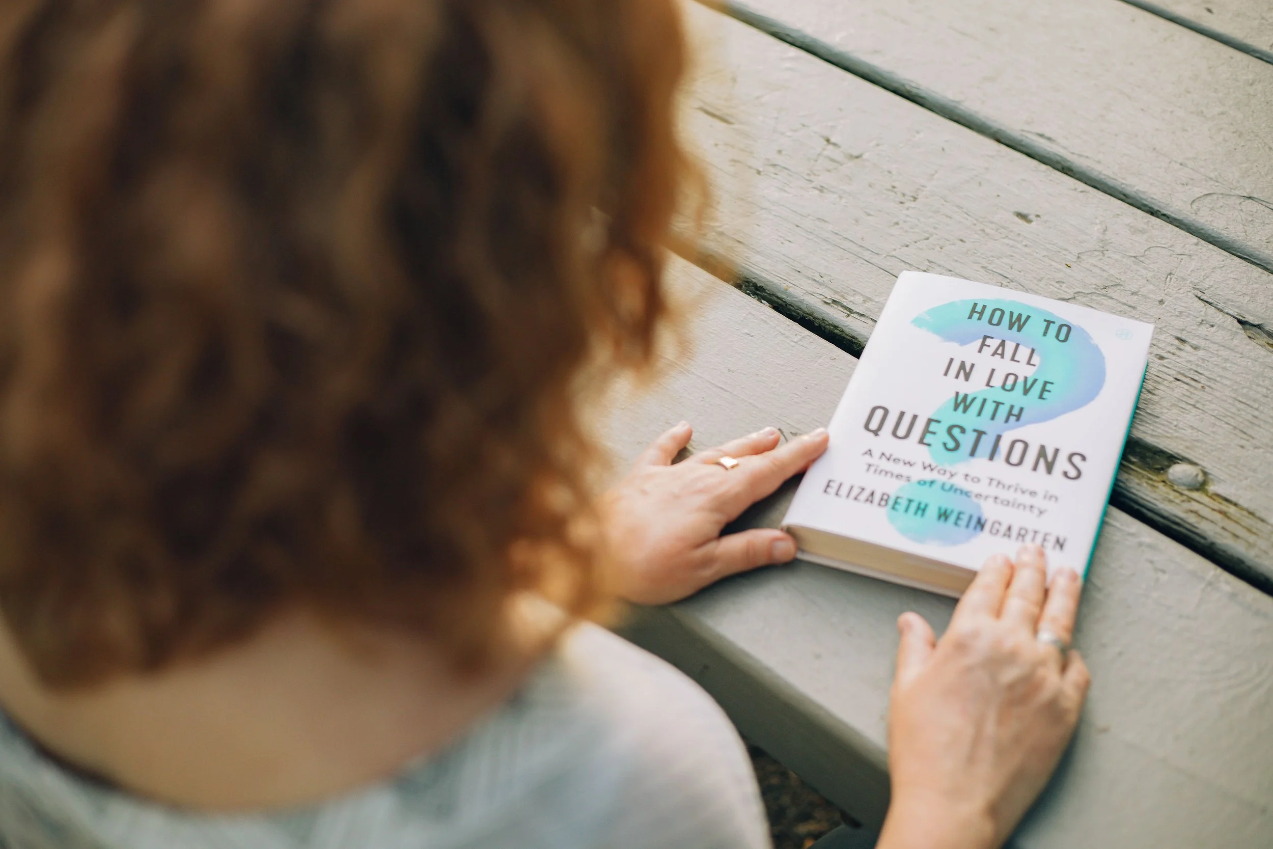 Photo of Jaye Matthews from behind sitting at a bench table with her hands on a book titled, "How to Fall in Love with Questions" by Elizabeth Weingarten