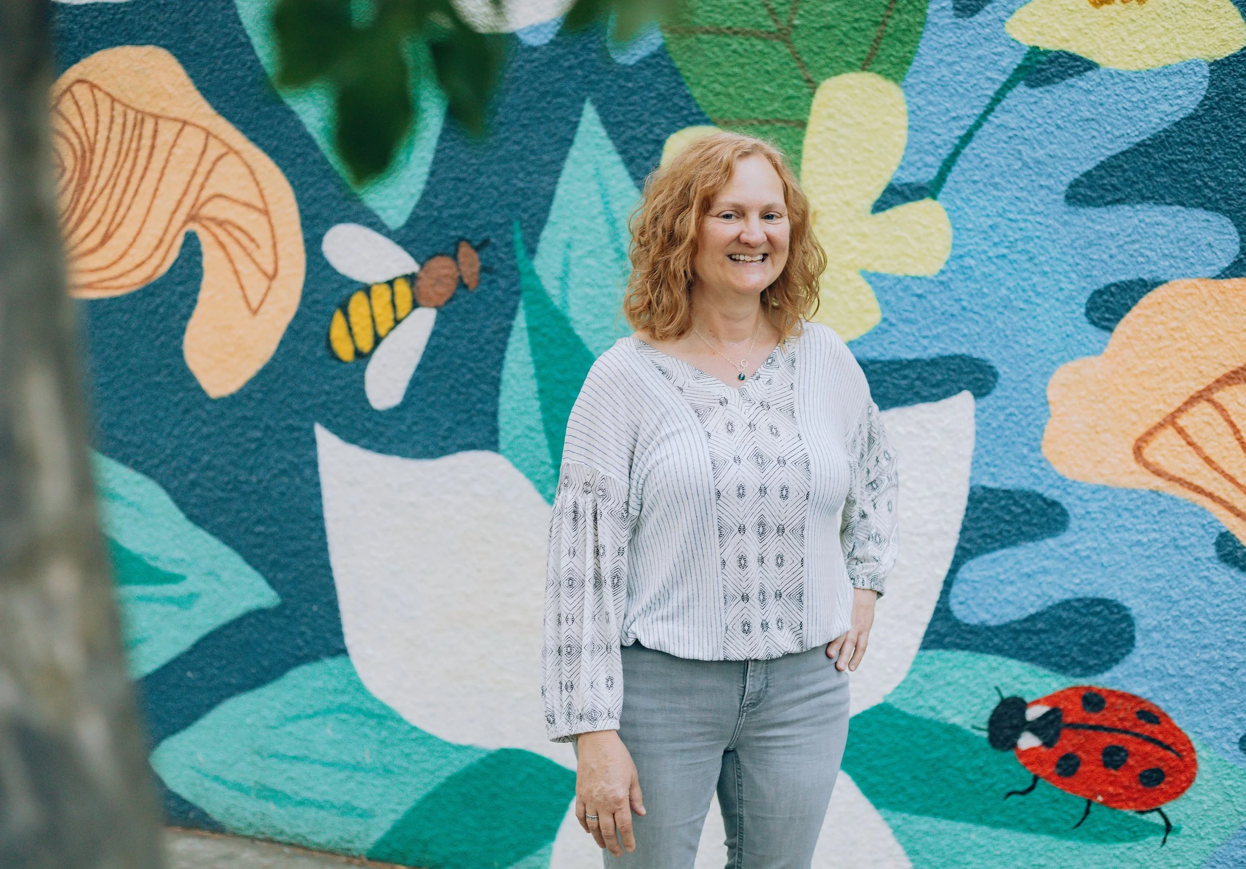 Photo of Jaye Matthews, a curly haired white woman smiling and standing in front of a blue, green, and white painted wall mural with a bee and a lady bug