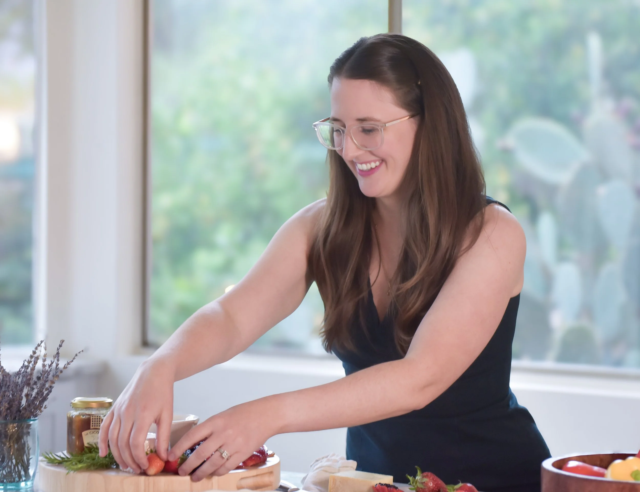 A woman with brown hair arranging fruit on a cutting board, smiling, with a window behind her