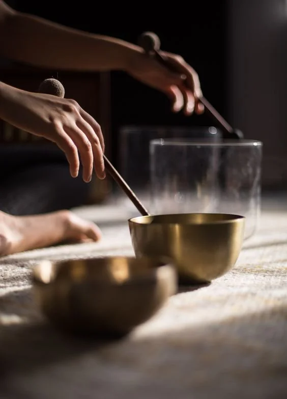 Two people are playing singing bowls with mallets on a wooden surface, with a glass container in the background, in a dimly lit setting.