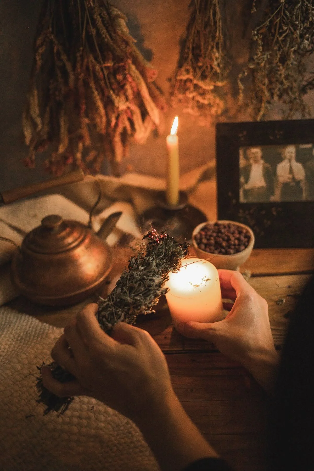 Hands placing a lit candle on a wooden table with dried herbs, a teapot, a bowl of herbs, a lit candle, dried herbs hanging on the wall, a framed picture of two people, and a small bowl of seeds.