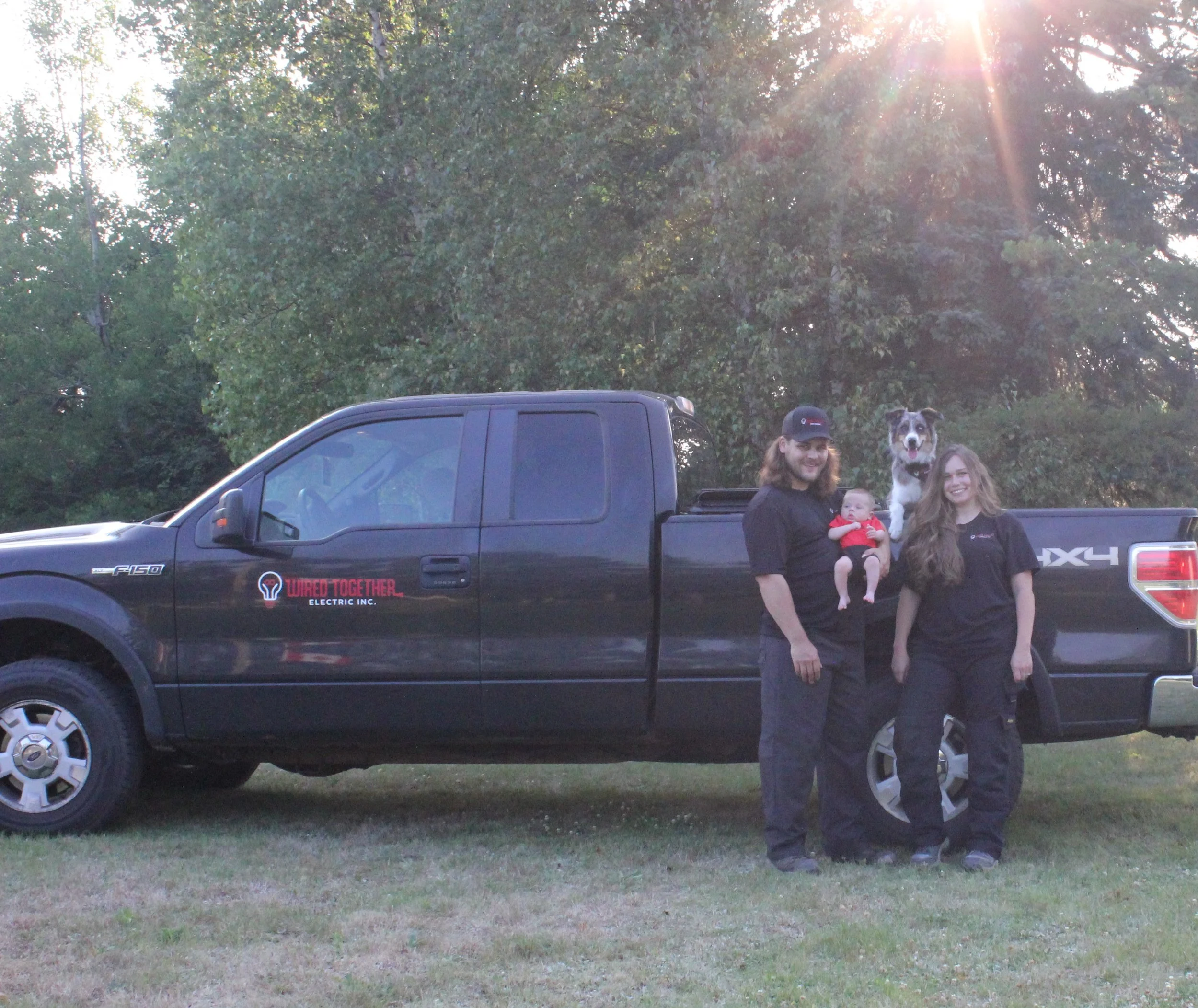 Two people, a man and a woman, stand in front of a black Ford F-150 pickup truck with a dog inside the truck bed. The man is holding a baby. The background has trees and sunlight.
