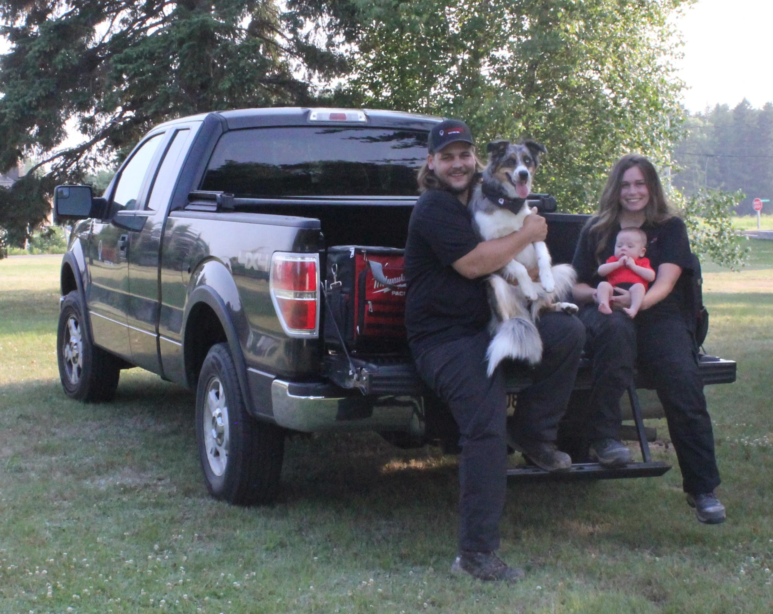 Two smiling people, a man and a woman, sitting on the back of a black pickup truck outdoors in a park, with a man holding a dog and a woman holding a baby, with trees and a grassy field in the background.