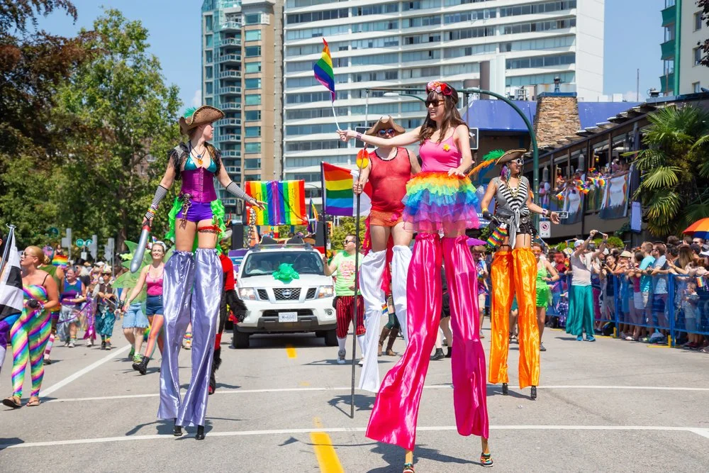 Parade with people on stilts dressed in colorful rainbows, rainbow flags, and rainbow-themed outfits on city street with tall buildings and crowd watching.