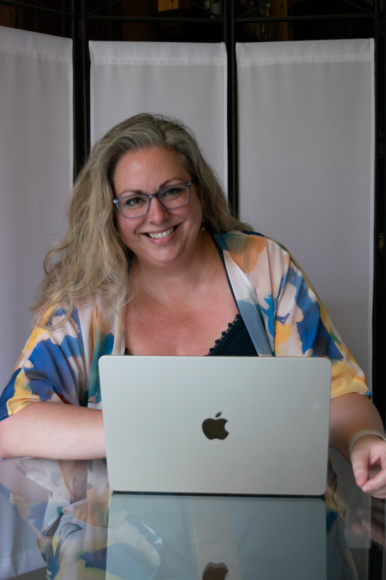 Photograph of Sarah Cozart, a blond woman with glasses, sitting at a glass table with an Apple laptop. She is wearing a blue and yellow loose sheer sweater over a navy blue dress.
