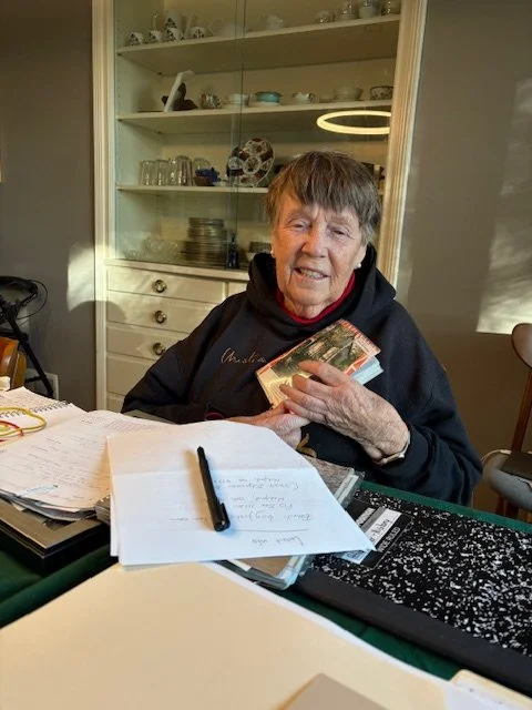 An Backstory senior client, holding documents from her life history, sitting at a table with papers, a pen, and a notebook, holding a deck of playing cards and smiling.