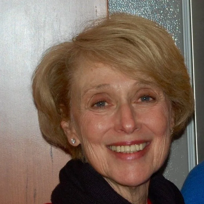 A smiling Backstory client who is an older woman with short, blong hair, wearing earrings and dark top, standing near a window.