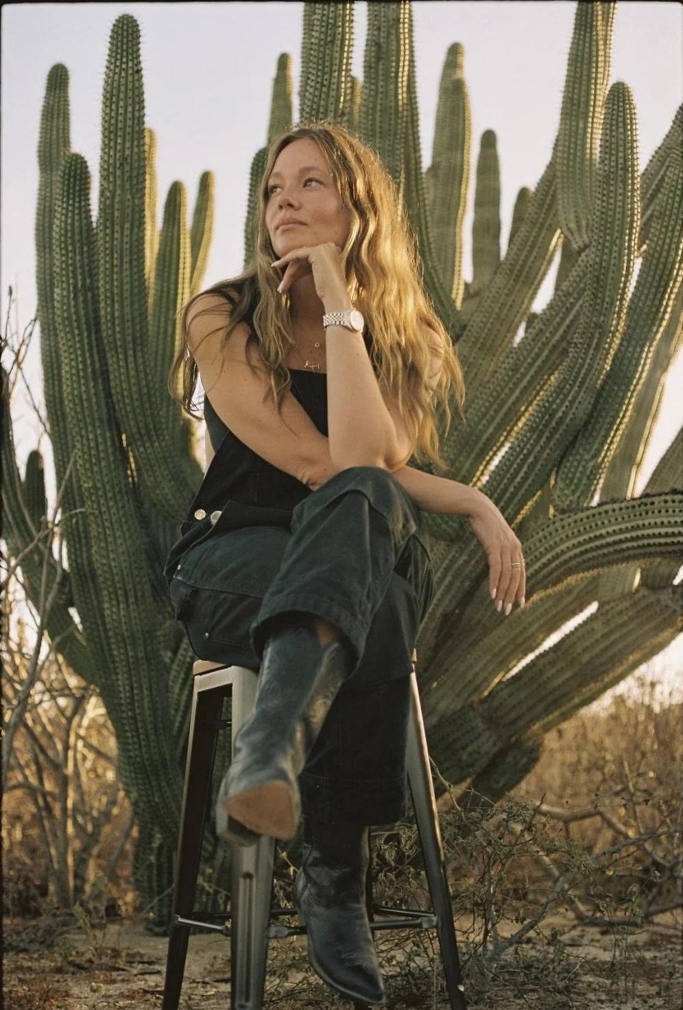 A woman sitting on a stool in front of large cactus plants, resting her chin on her hand, wearing black clothing and boots, with a desert landscape in the background.