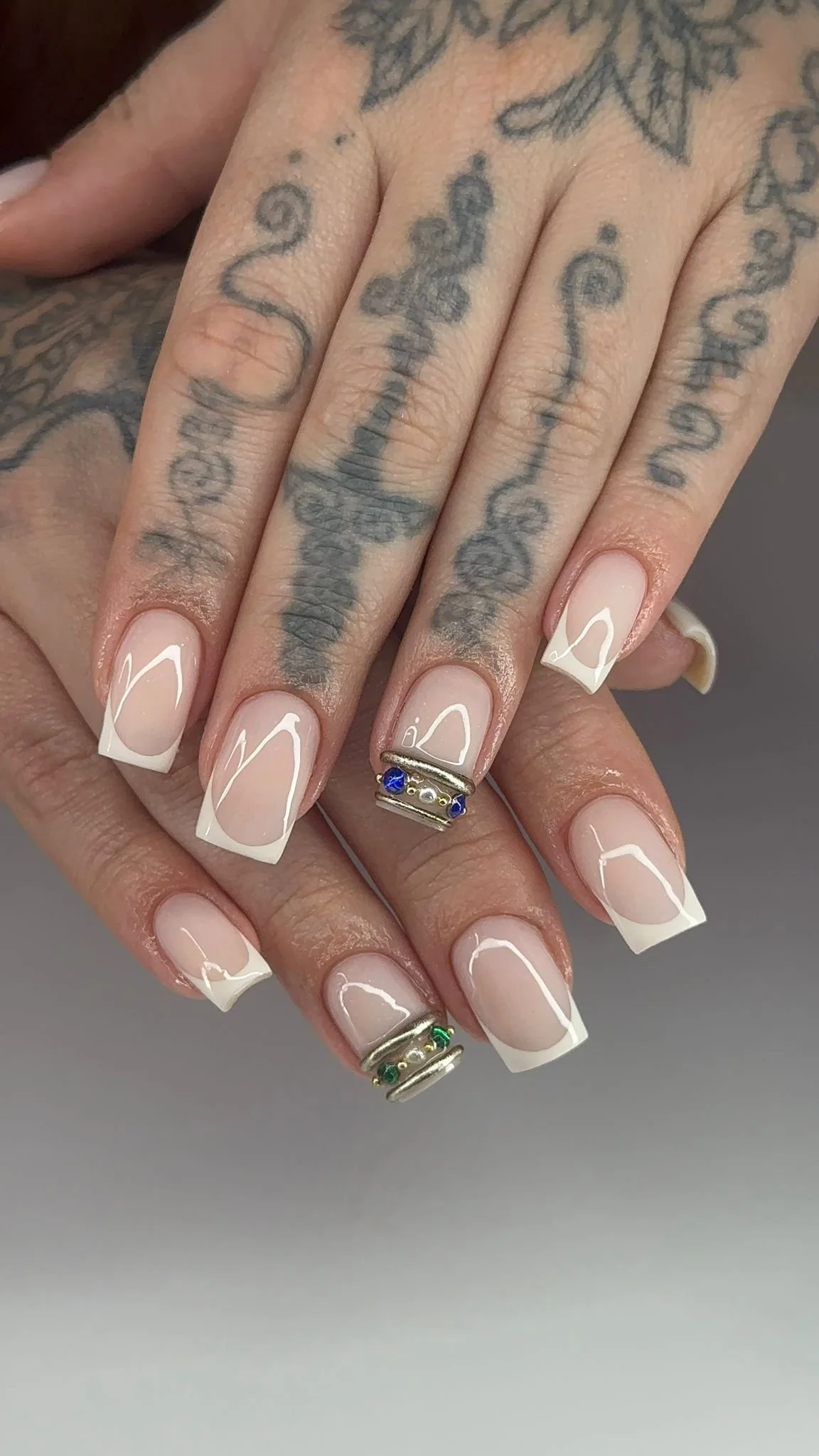 Close-up of two hands with manicured nails and tattoos on fingers and back of hands, featuring rings with blue and green stones.