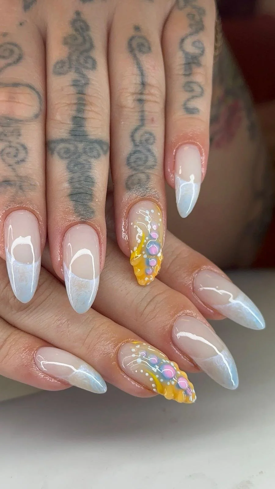 Nail art with white tips and colorful designs on ring fingers, tattoos on fingers and hand, skin with visible tattoos.
