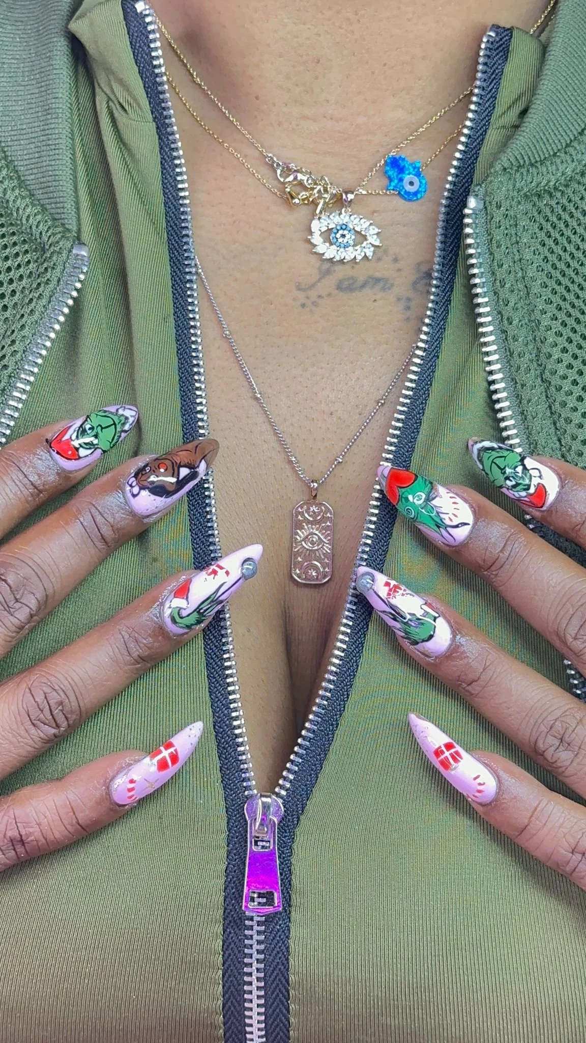 Close-up of a person's chest and hands, showing layered necklaces, one with a pendant featuring an eye design, and colorful, detailed nail art with various illustrations including faces and flags. The person is wearing a green zip-up jacket with a textured fabric.