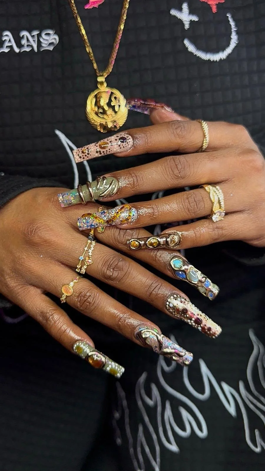 A person showcasing multiple rings on their fingers and decorated long nails, wearing a gold chain necklace, with a dark background.