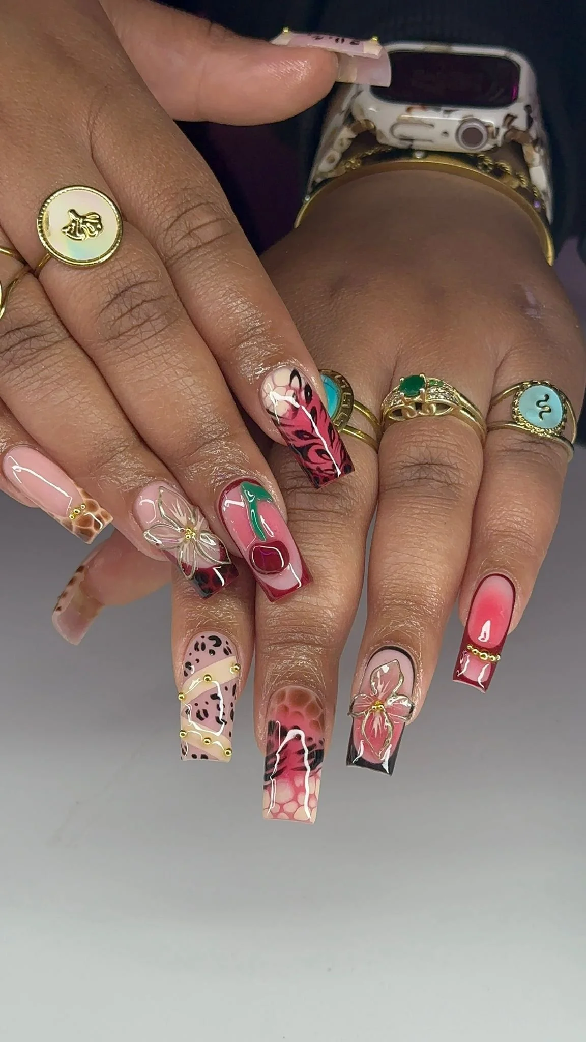 Close-up of a hand with decorated nails, rings, and a wristwatch. The nails are long, square-shaped, with colorful, intricate designs including flowers, animal prints, and abstract patterns.