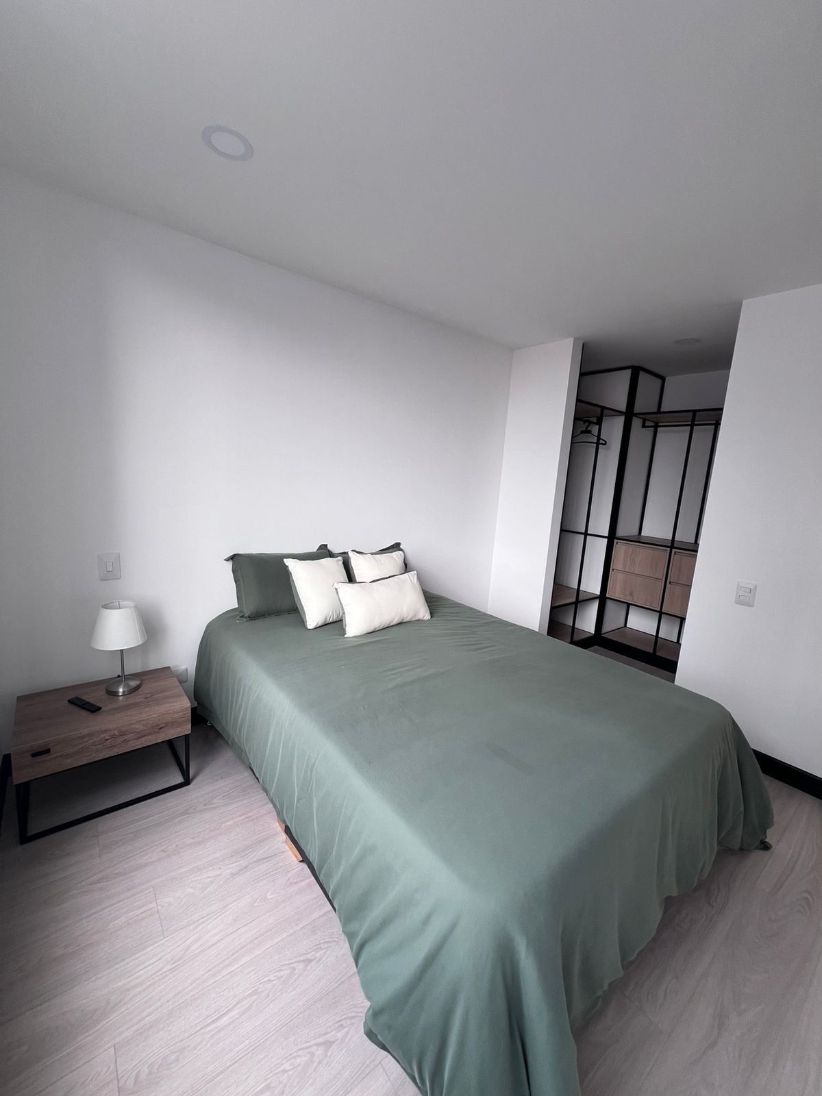 Habitación con cama doble, almohadas blancas y marrones, ventanas grandes con vista a edificios y árboles, y suelo de madera.