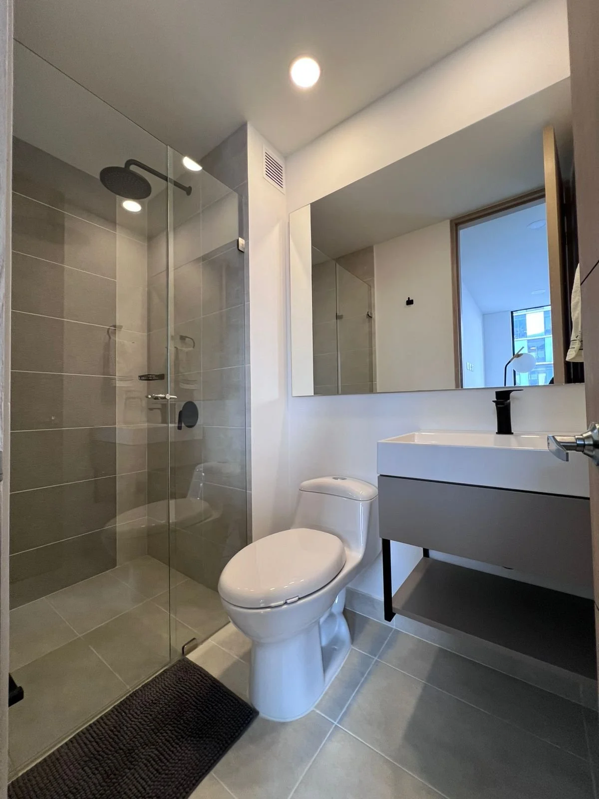 Habitación con ventanas panorámicas, cama con sábanas blancas y cobertor marrón, almohadas blancas y marrón, suelo de madera, paredes blancas, armario negro y TV en la esquina.