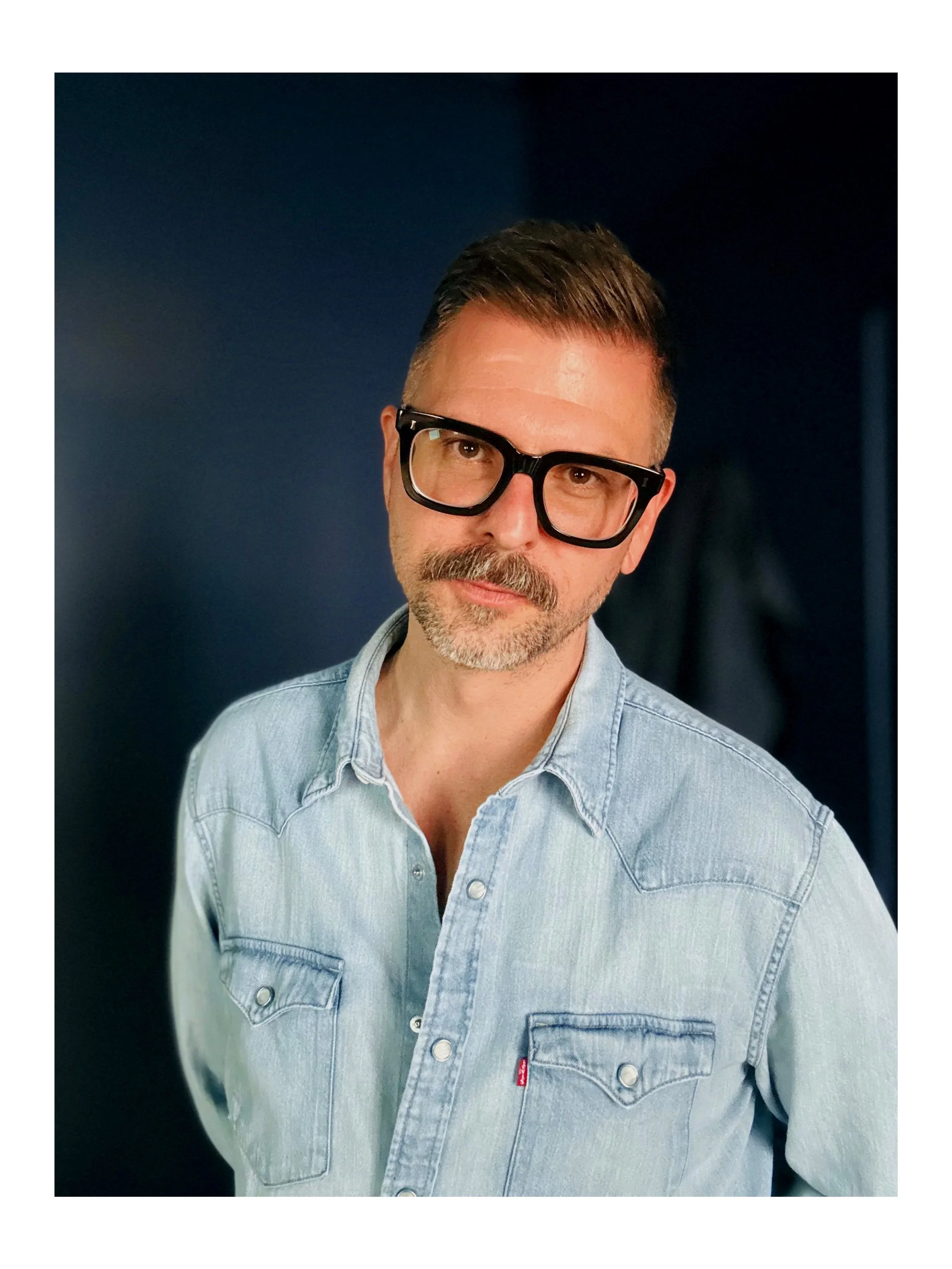 Man wearing glasses and a denim shirt, posing against a dark background.