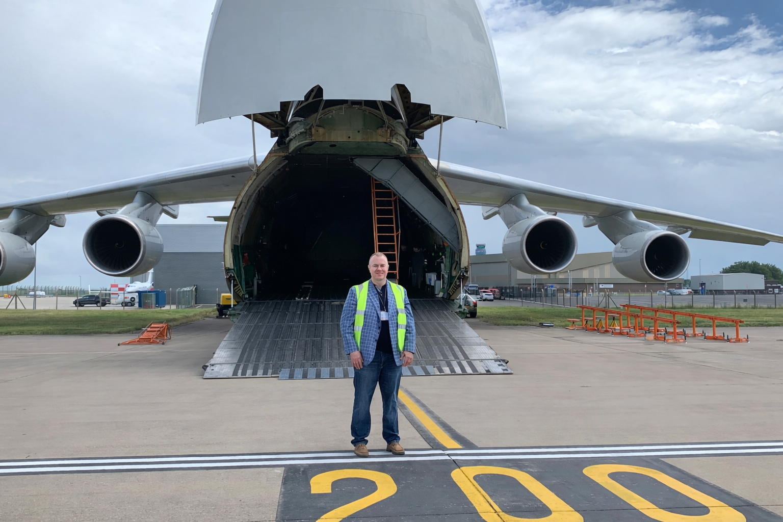 A man in a safety vest and casual clothing standing in front of a large cargo aircraft with an open rear cargo ramp at an airport.