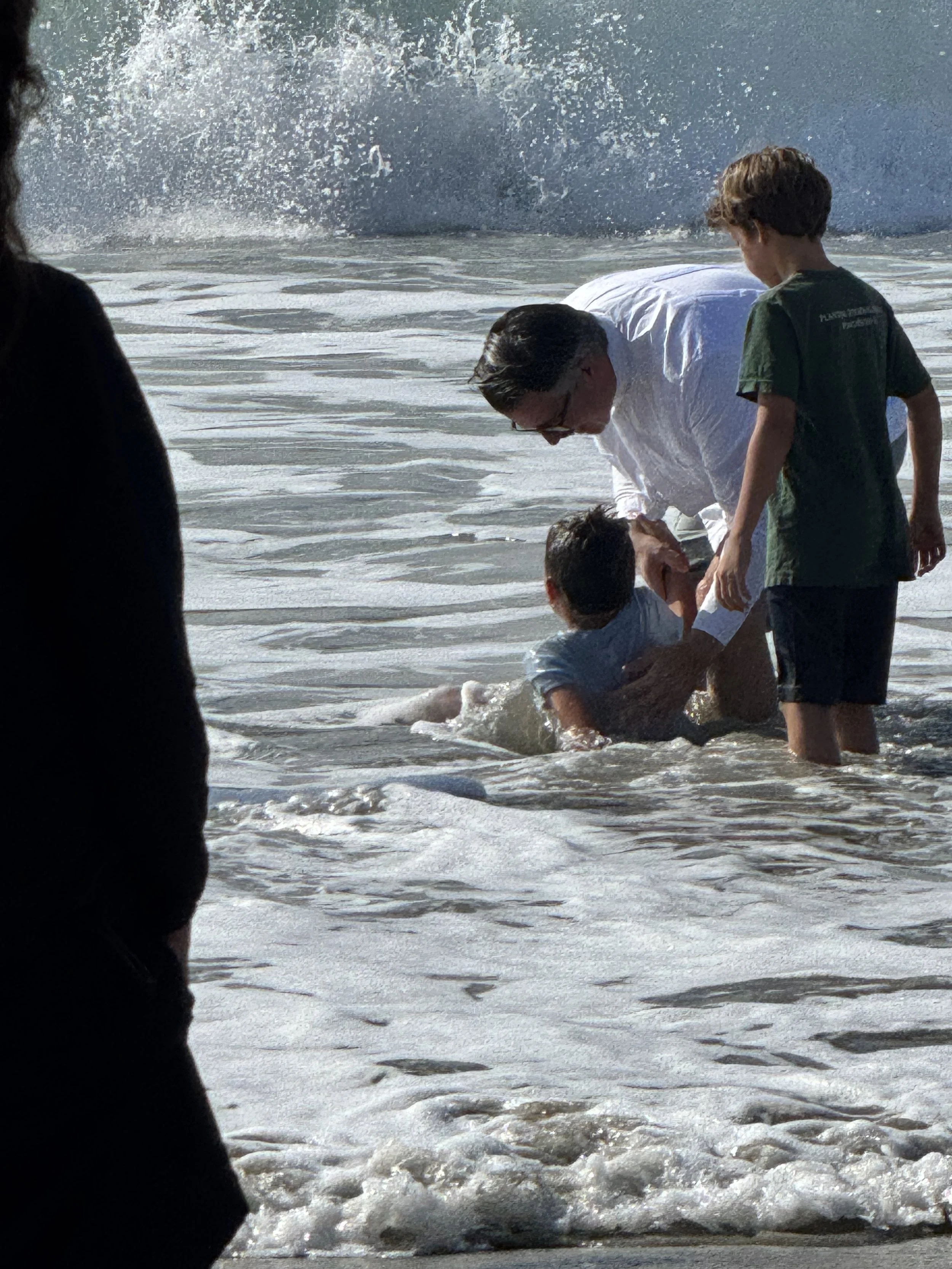 Pastor Daniel Hyde baptizing a children in the ocean and another child is a friend standing nearby, as a wave crashes in the background.
