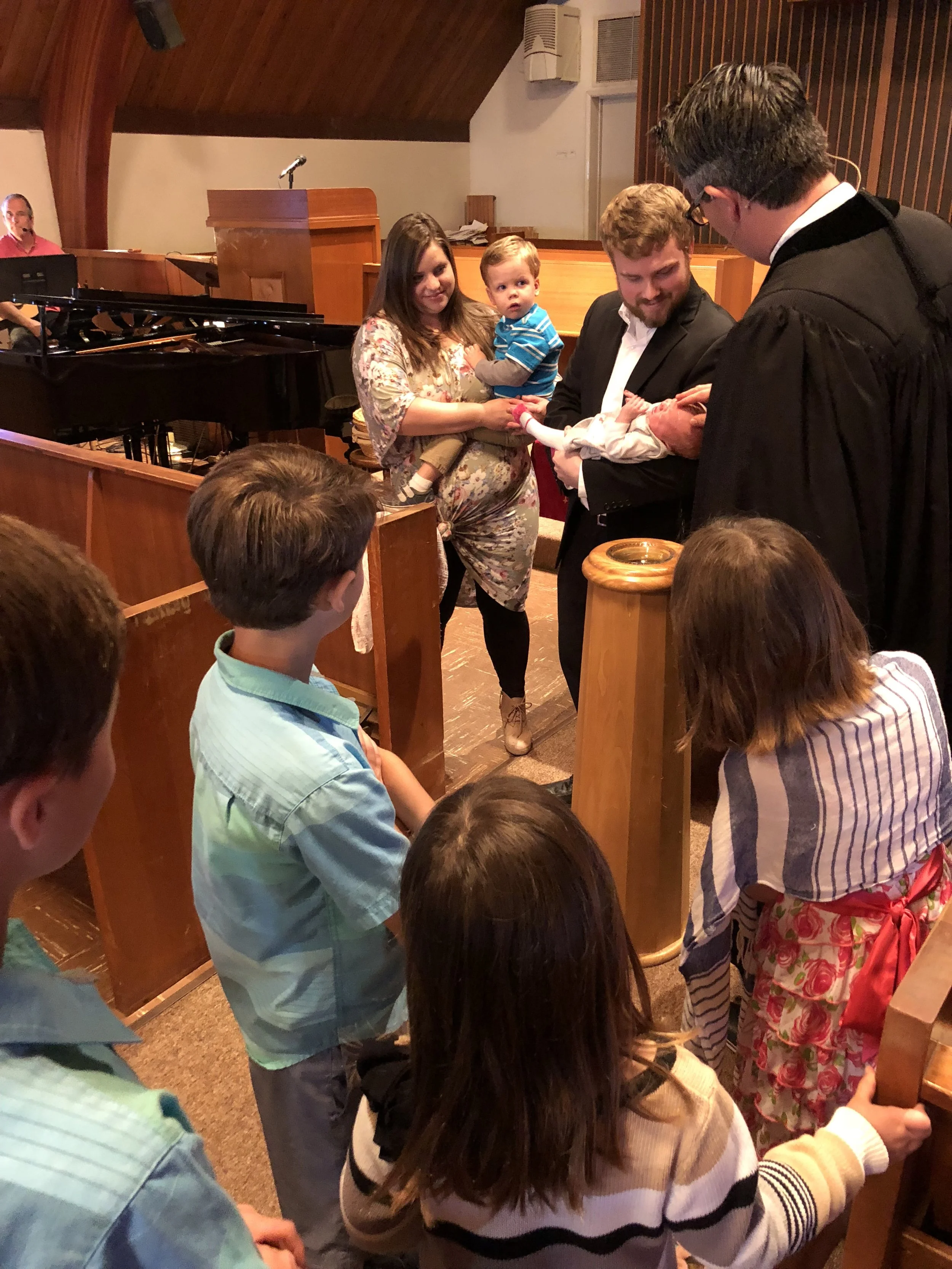 A baptism ceremony taking place in a church with pastor Daniel Hyde, a baby, and family members gathered around.