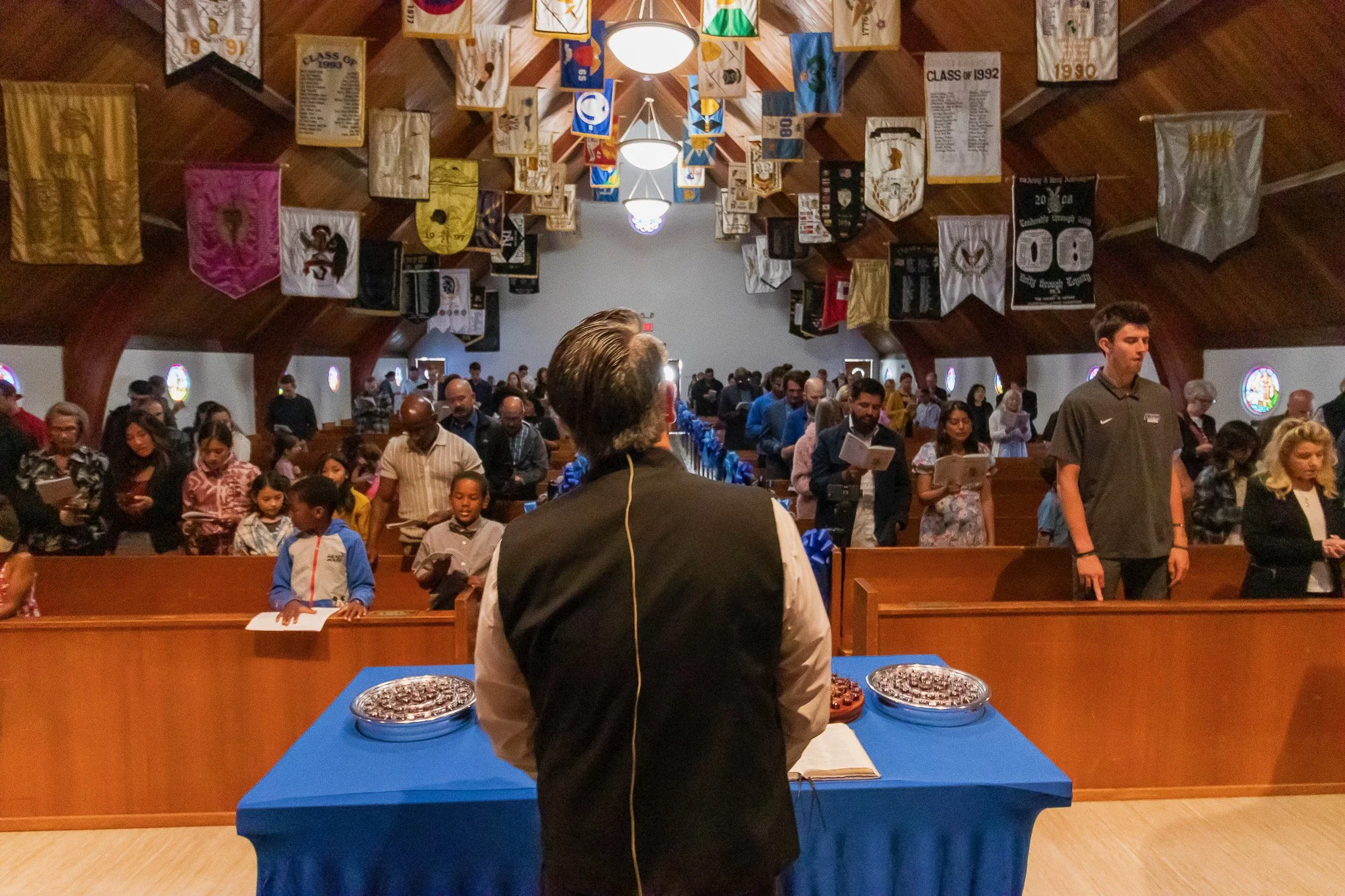 Inside the Chapel of the Army & Navy Academy where Oceanside URC meets. This event was its 25th anniversary celebration in May 2025. There are children and adults standing and singing with Pastor Danny inn front behind the Communion Table.