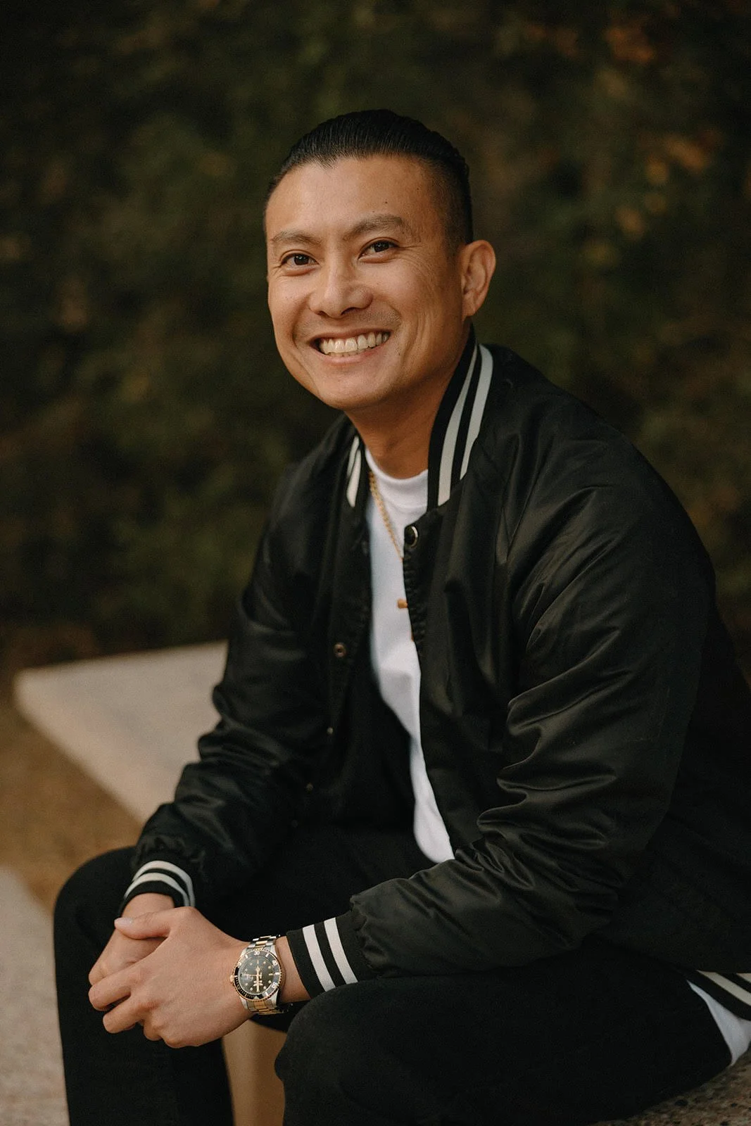 Kevin Mai, the Young Adults ministry coordinator at Oceanside URC, smiling in a black jacket, white shirt, and watch sitting on a bench outdoors with a blurred natural background.