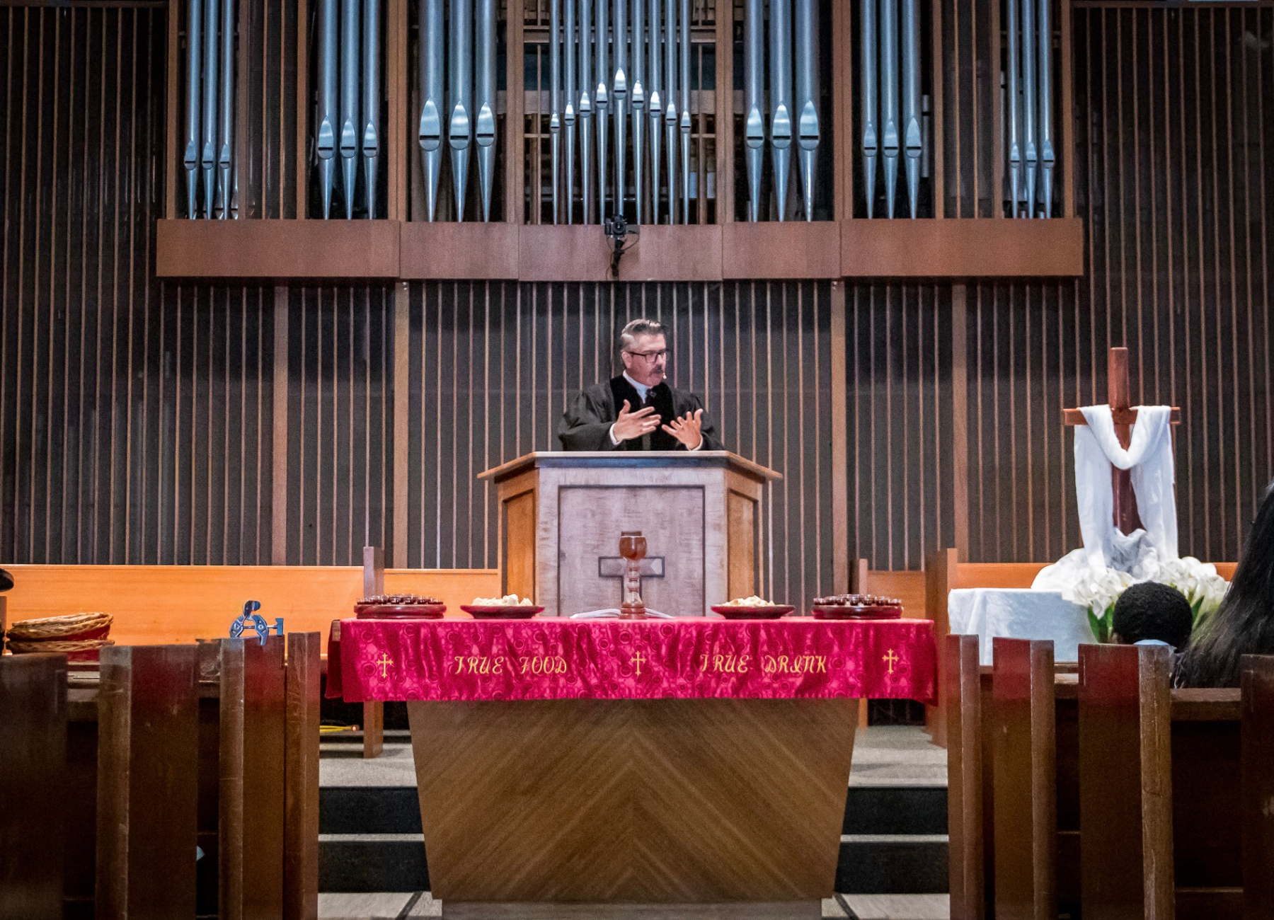 Dr. Daniel Hyde in black Genevan preaching robe standing behind the pulpit at Oceanside United Reformed Church, with a large pipe organ in the background and a table with communion items in the foreground.