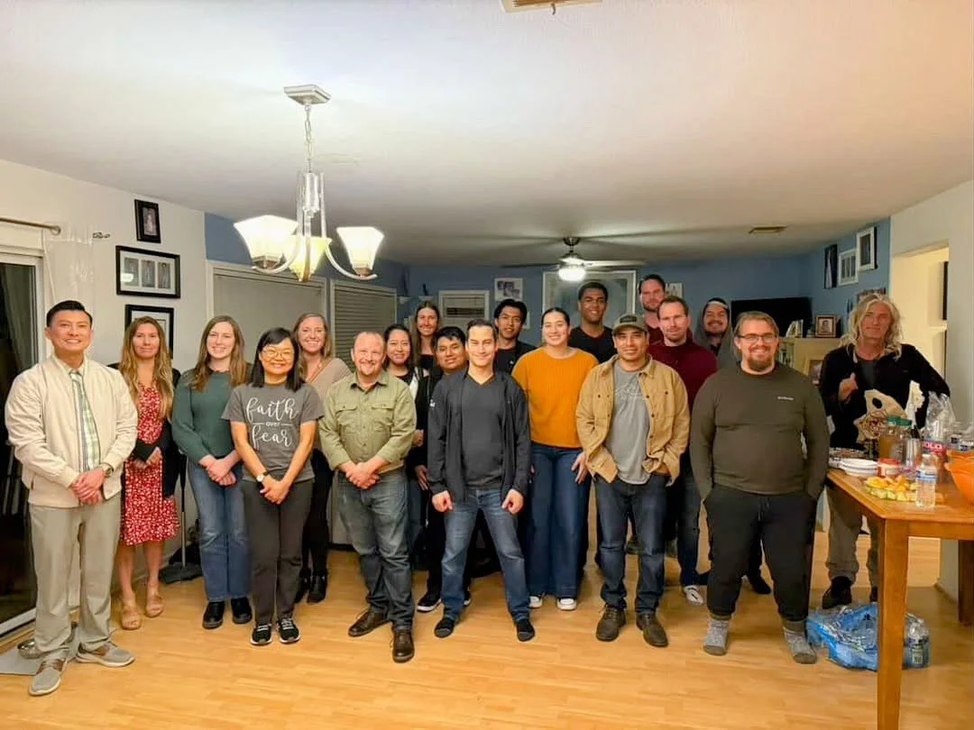 Group of 18 people gathered in a living room, standing and smiling for a photo.