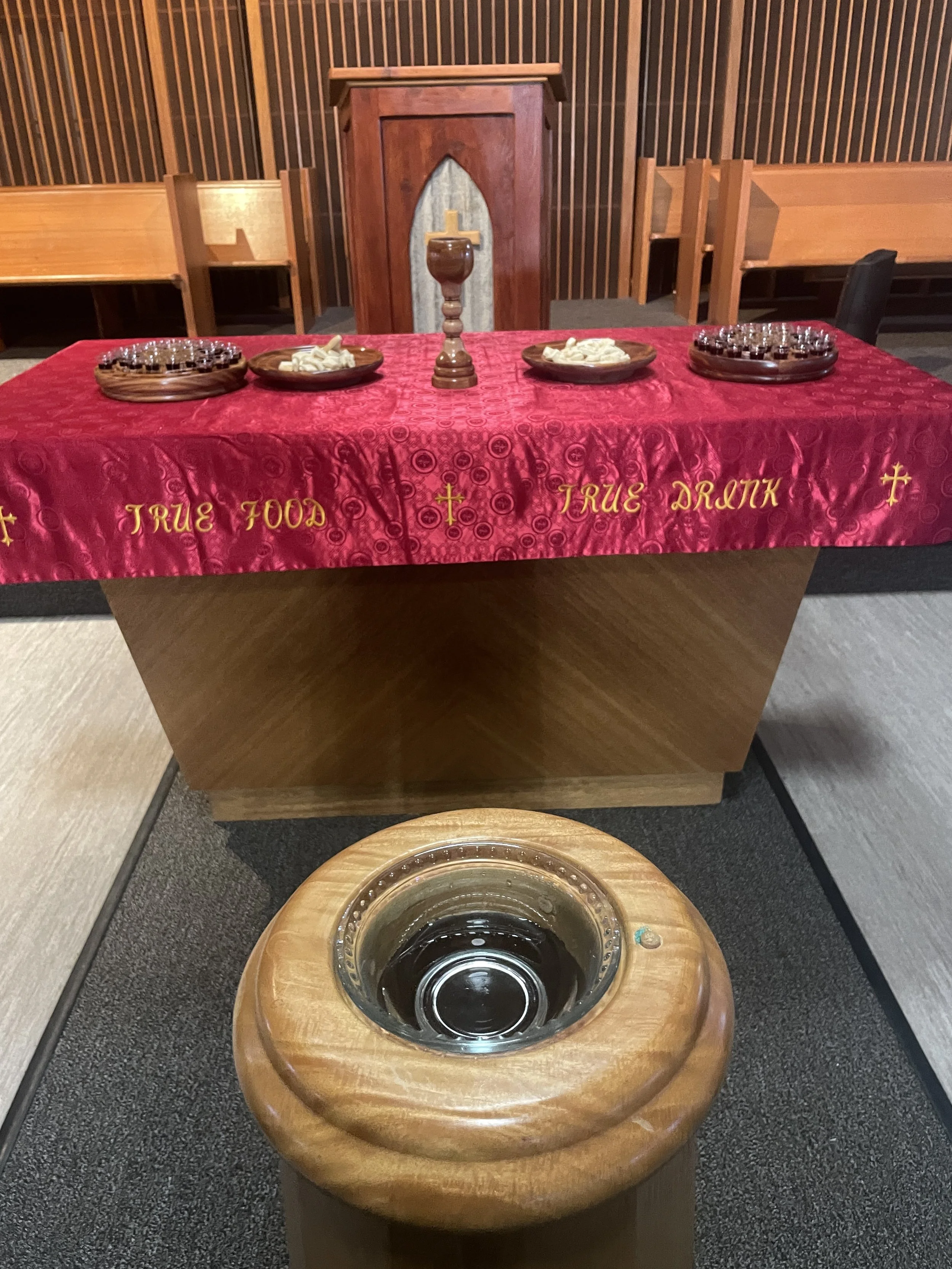 A communion table covered with a red cloth that reads 'True Food' and 'True Drink', and contains a cup, plates with bread, and trays with small cups, in a church setting.