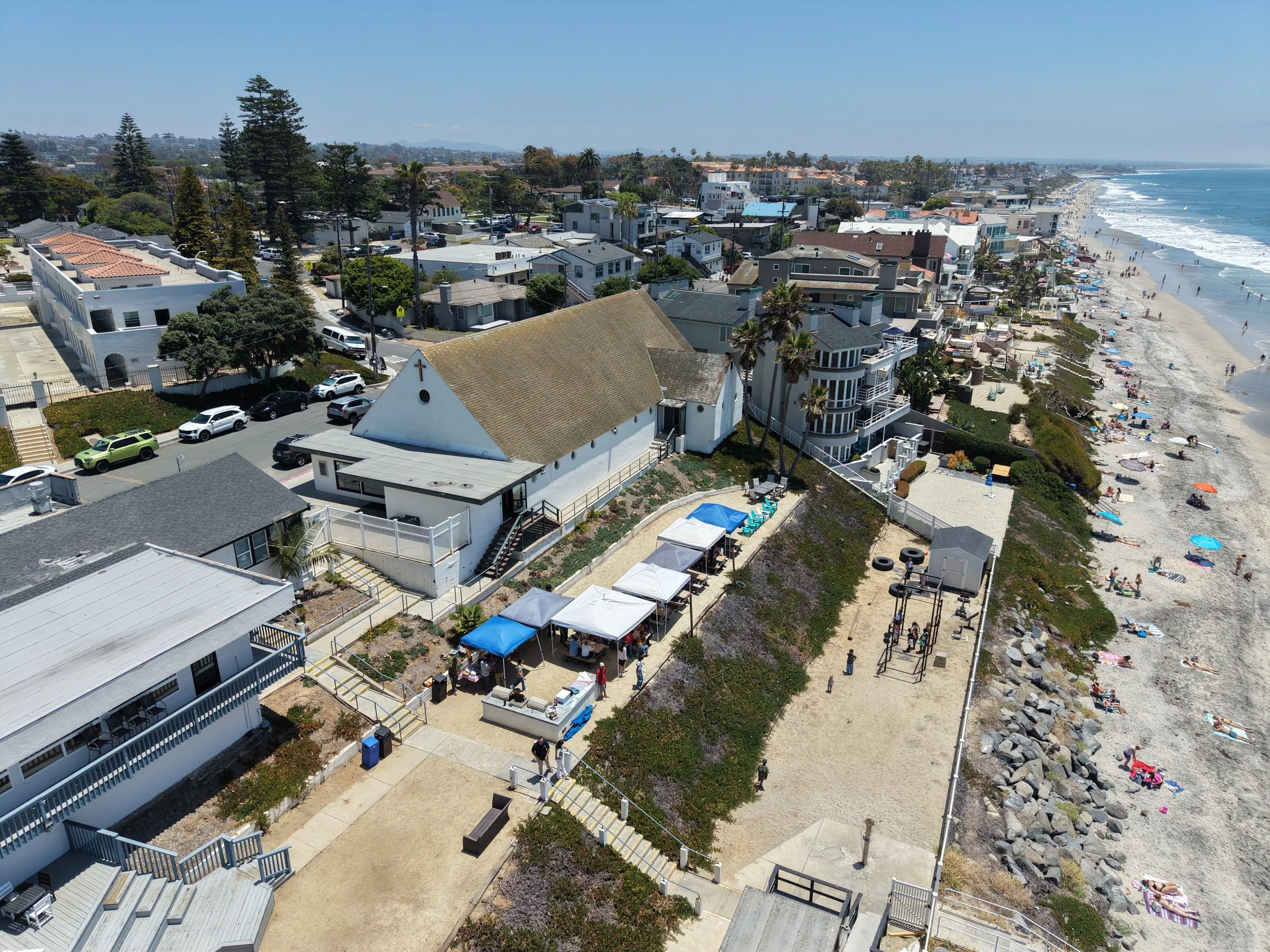 Aerial view of a the Chapel of the Army & Navy Academy where the Oceanside United Reformed Church meets along the coast of Carlsbad, with neighborhood houses and streets near a crowded beach with umbrellas and people in the ocean.