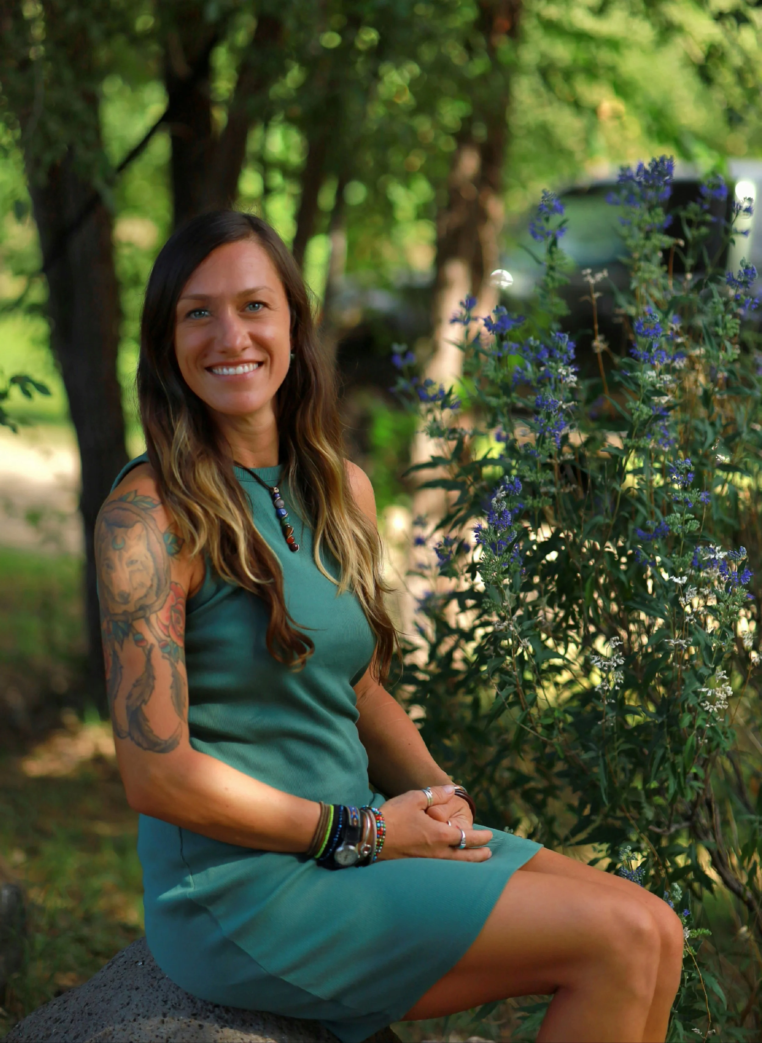 A woman with long wavy hair, wearing a green dress, sitting outdoors on a rock, smiling at the camera, with tattoos on her arm, surrounded by greenery and purple flowers.