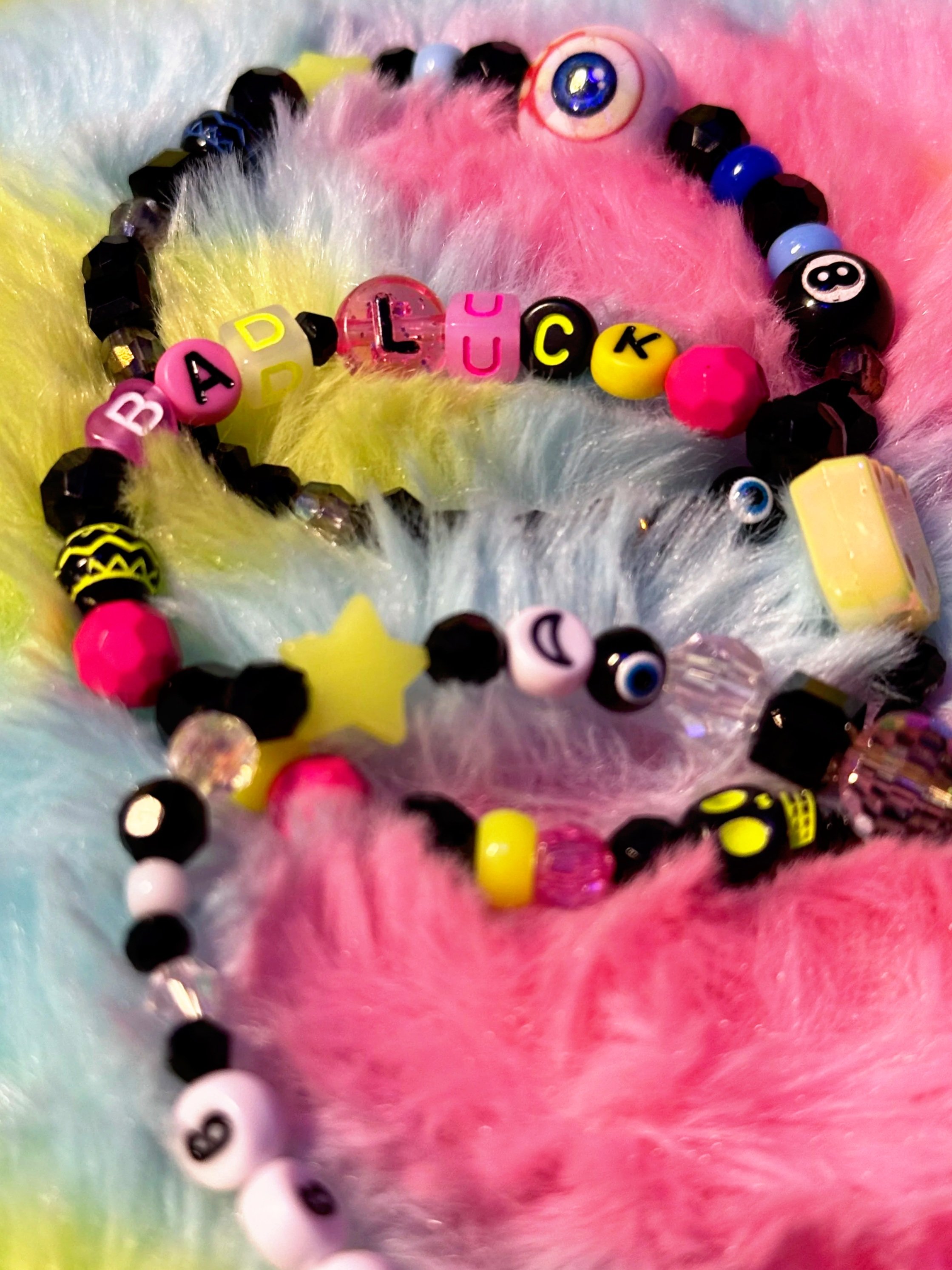 Colorful beaded bracelets with letters spelling "BAD LUCK" and various eye and star charms, resting on pastel-colored fluffy fabric.