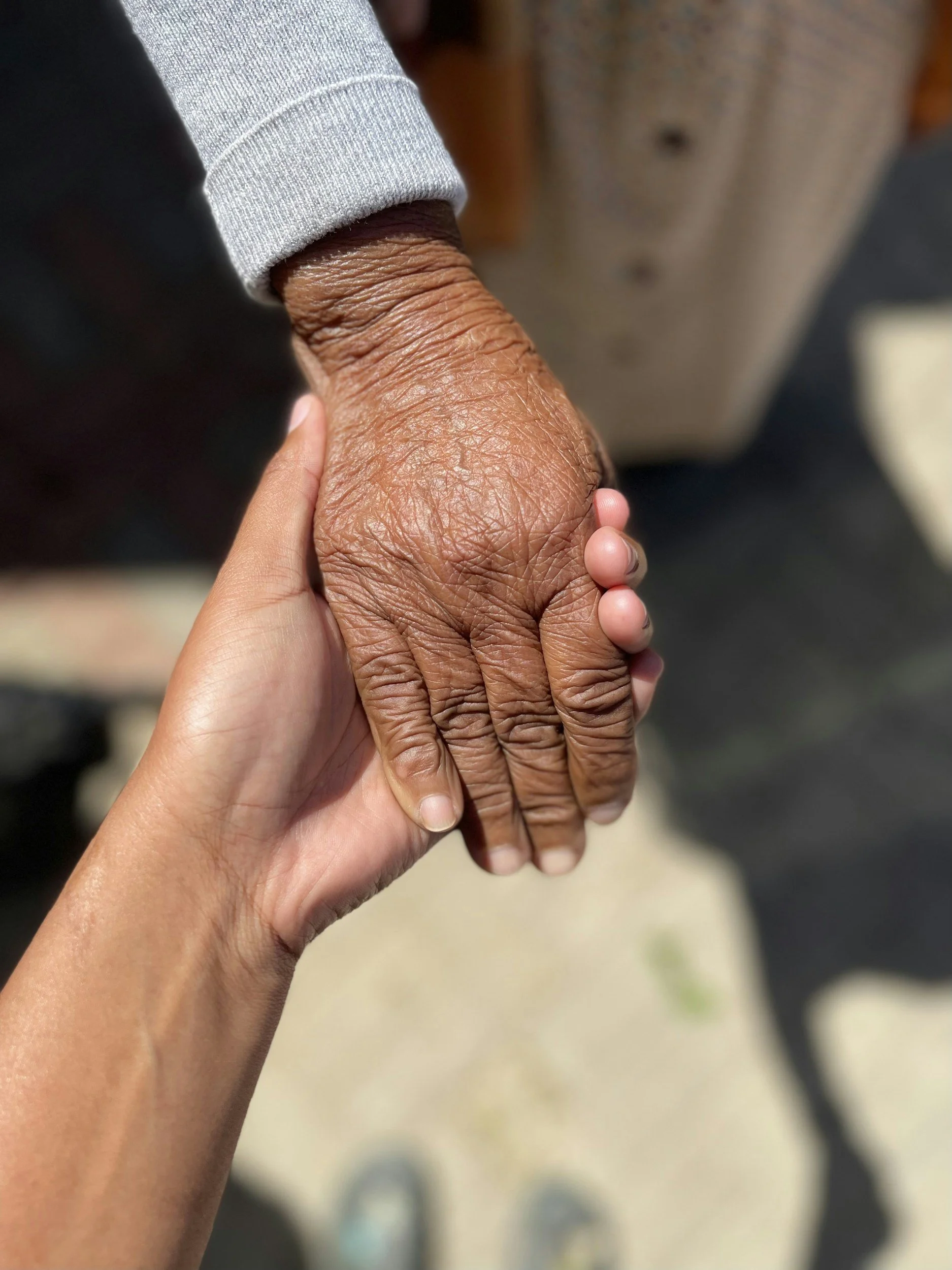 A human hand holding an elderly person's wrinkled hand in an outdoor setting.
