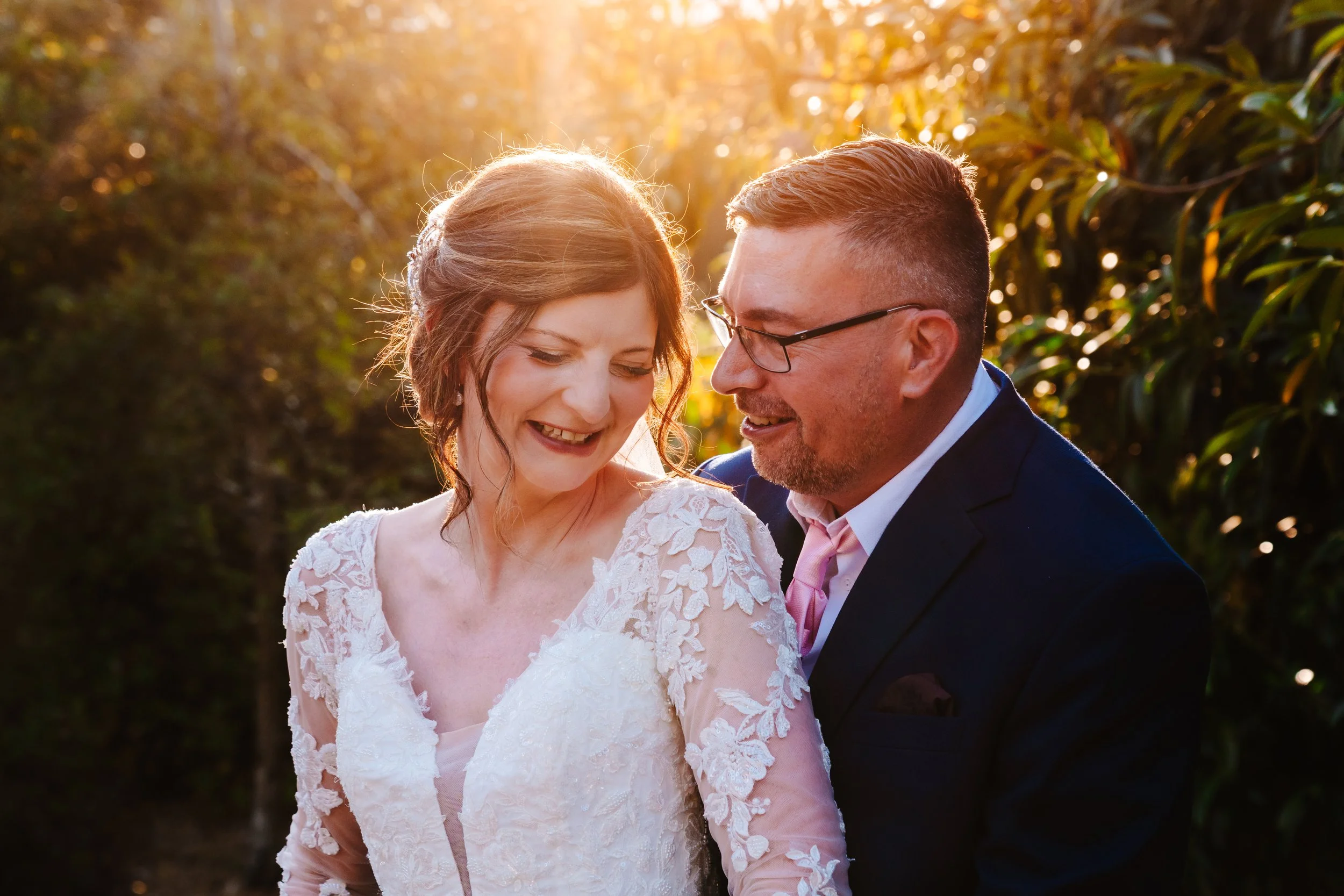 A bride and groom are smiling and looking at each other outdoors during sunset. The bride is wearing a white lace wedding dress, and the groom is wearing a dark suit with a pink tie. The background is filled with trees and sunlight.