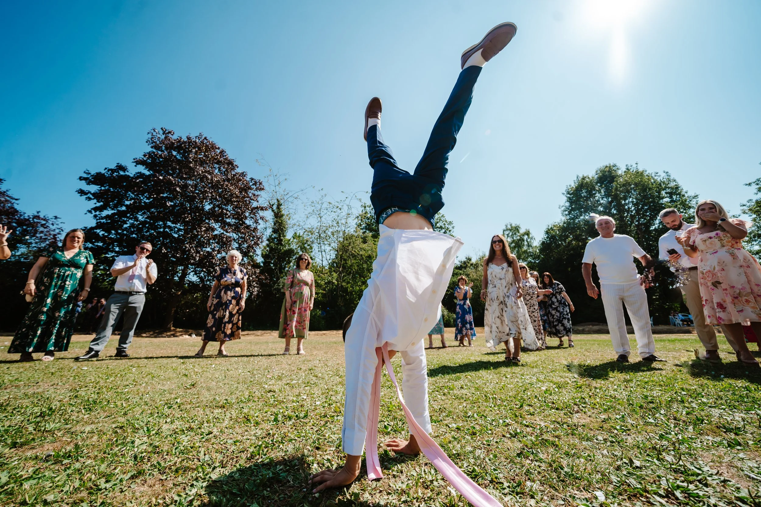 Person in formal attire upside down, participating in a limbo game during an outdoor gathering, with a group of people watching and smiling under a clear blue sky.