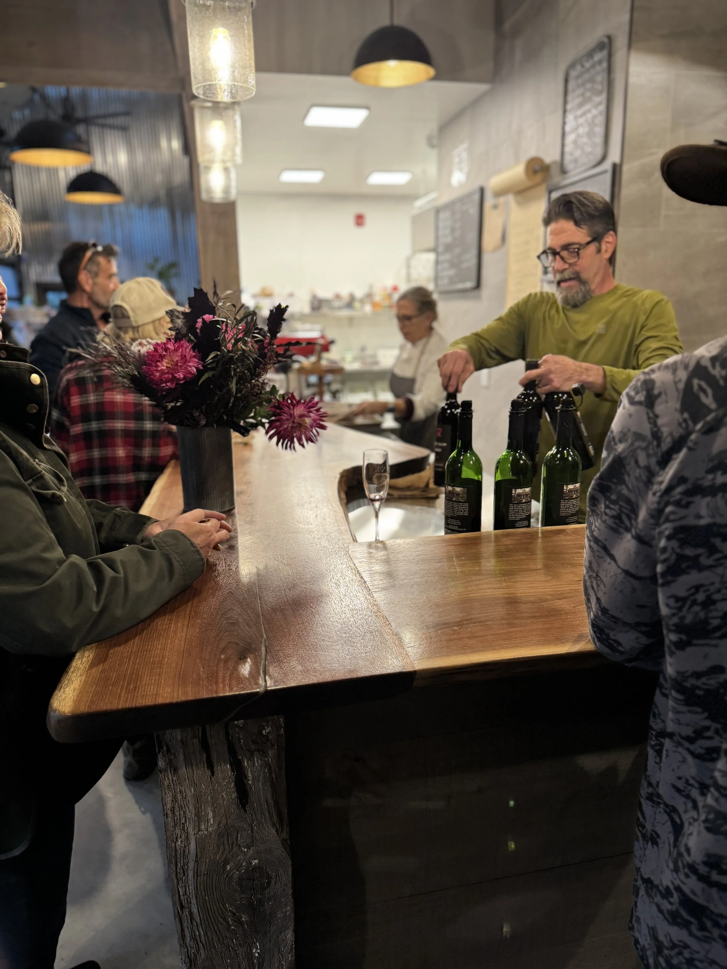 People seated at a wooden bar inside a restaurant, with drinks and a vase of pink and purple flowers on the bar. A bartender in a green shirt is opening bottles of wine or beer, while other staff and customers are visible in the background.