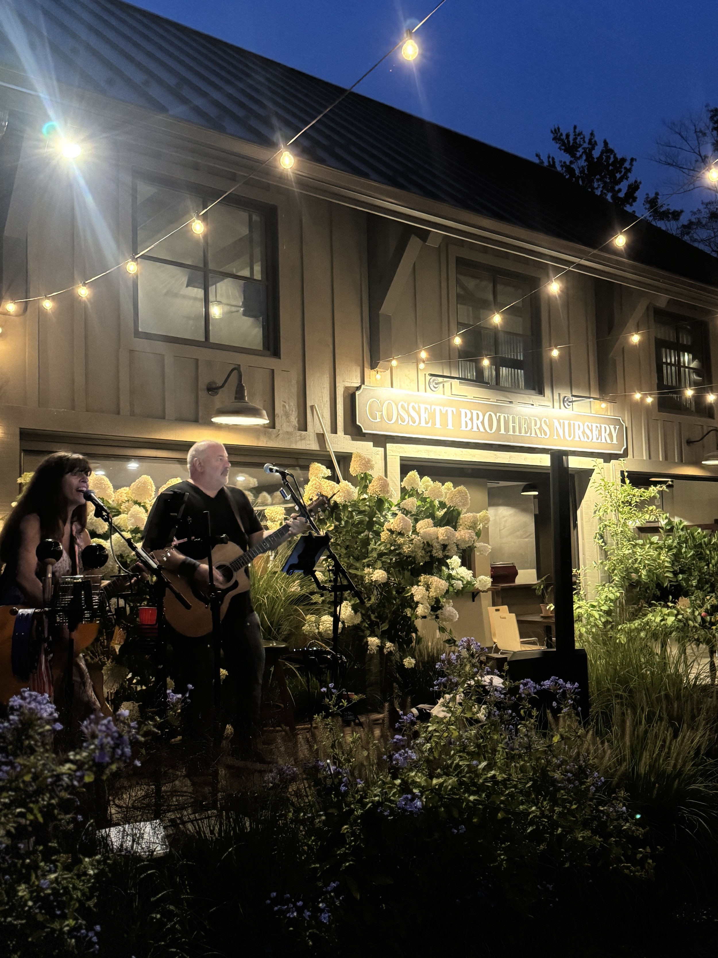 Two musicians performing with guitars and microphones outside Gossett Brothers Nursery at night, decorated with string lights and surrounded by white and purple flowers.