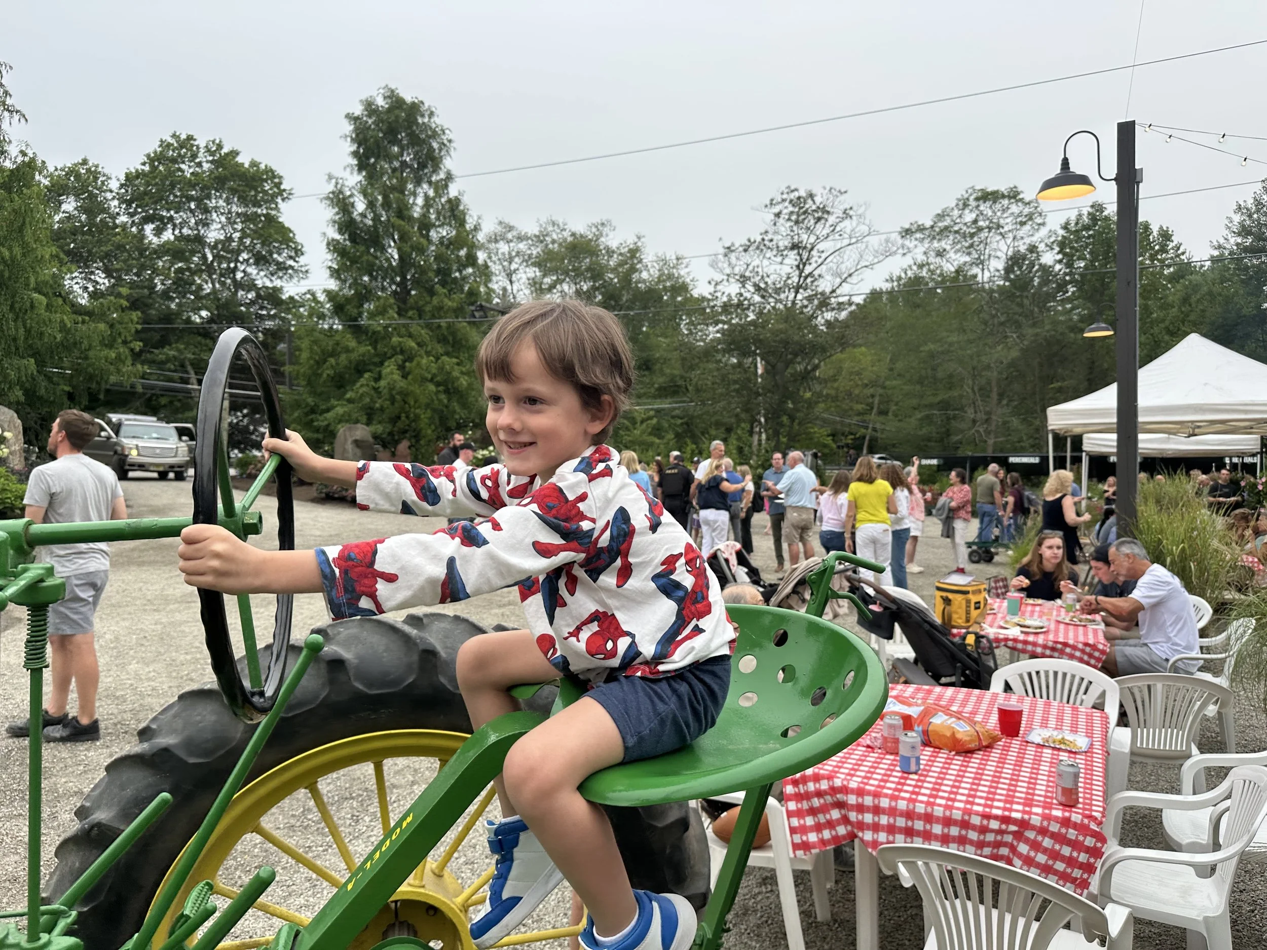 A young boy wearing a Spiderman shirt, shorts, and blue sneakers, smiling while sitting on a green tractor ride during an outdoor gathering with people in the background, some sitting at tables with red and white checkered tablecloths.