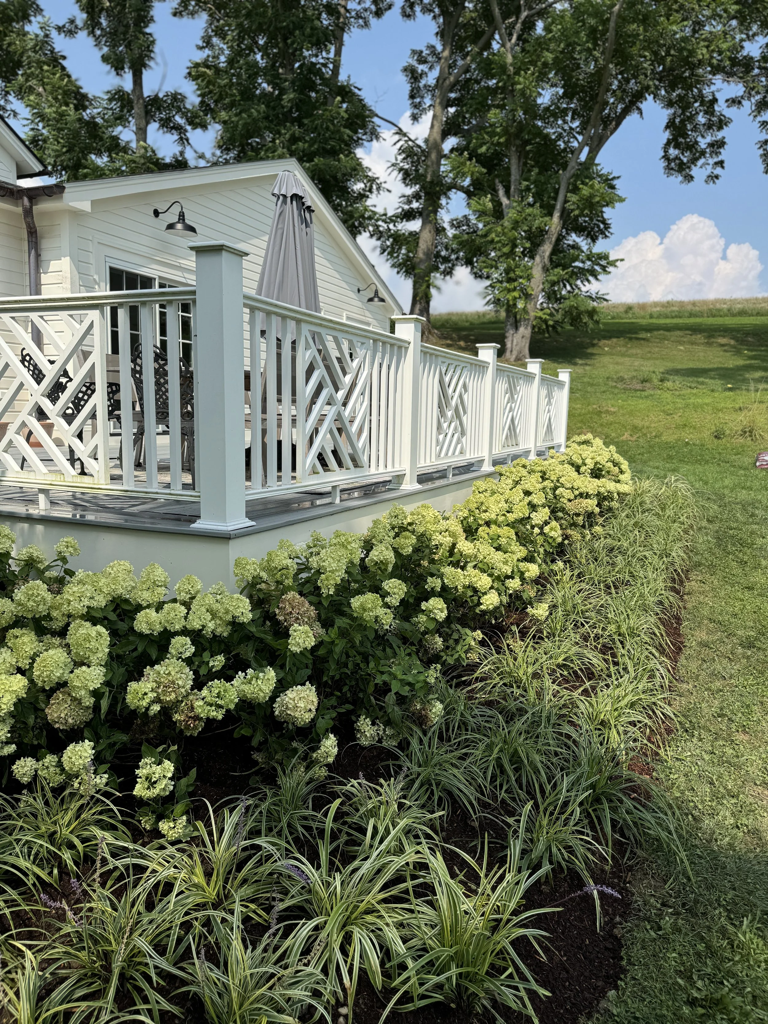 White house with a porch, white railing, and outdoor furniture, surrounded by flowering shrubs and green grass, with trees and blue sky in the background.