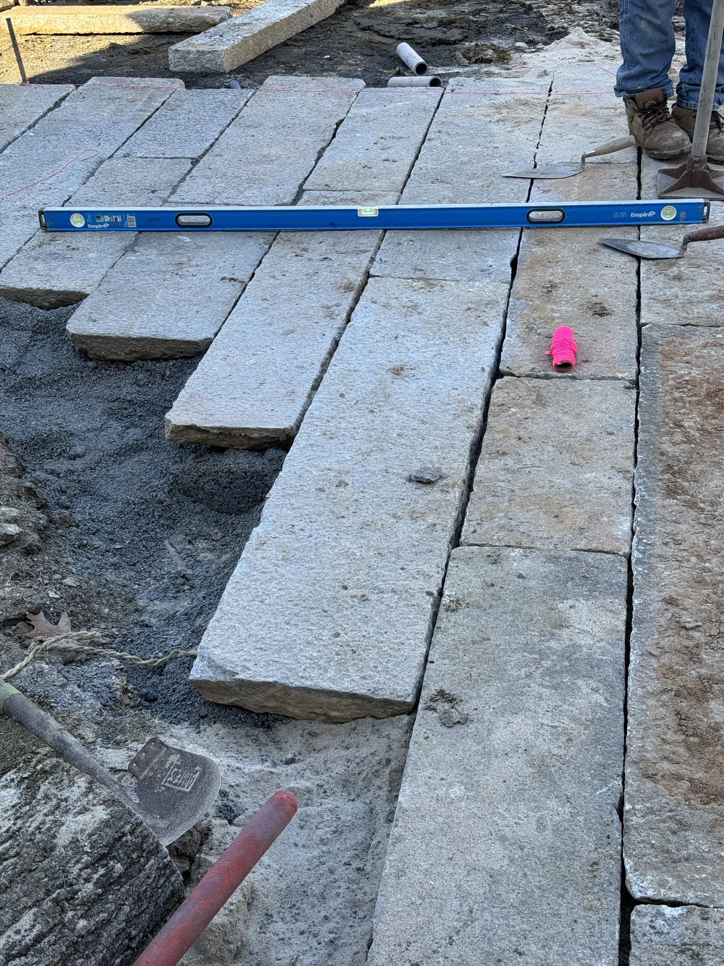 Construction site with large stone pavers arranged in a pathway, a level tool on top, pink string, and construction worker wearing brown boots and blue jeans.