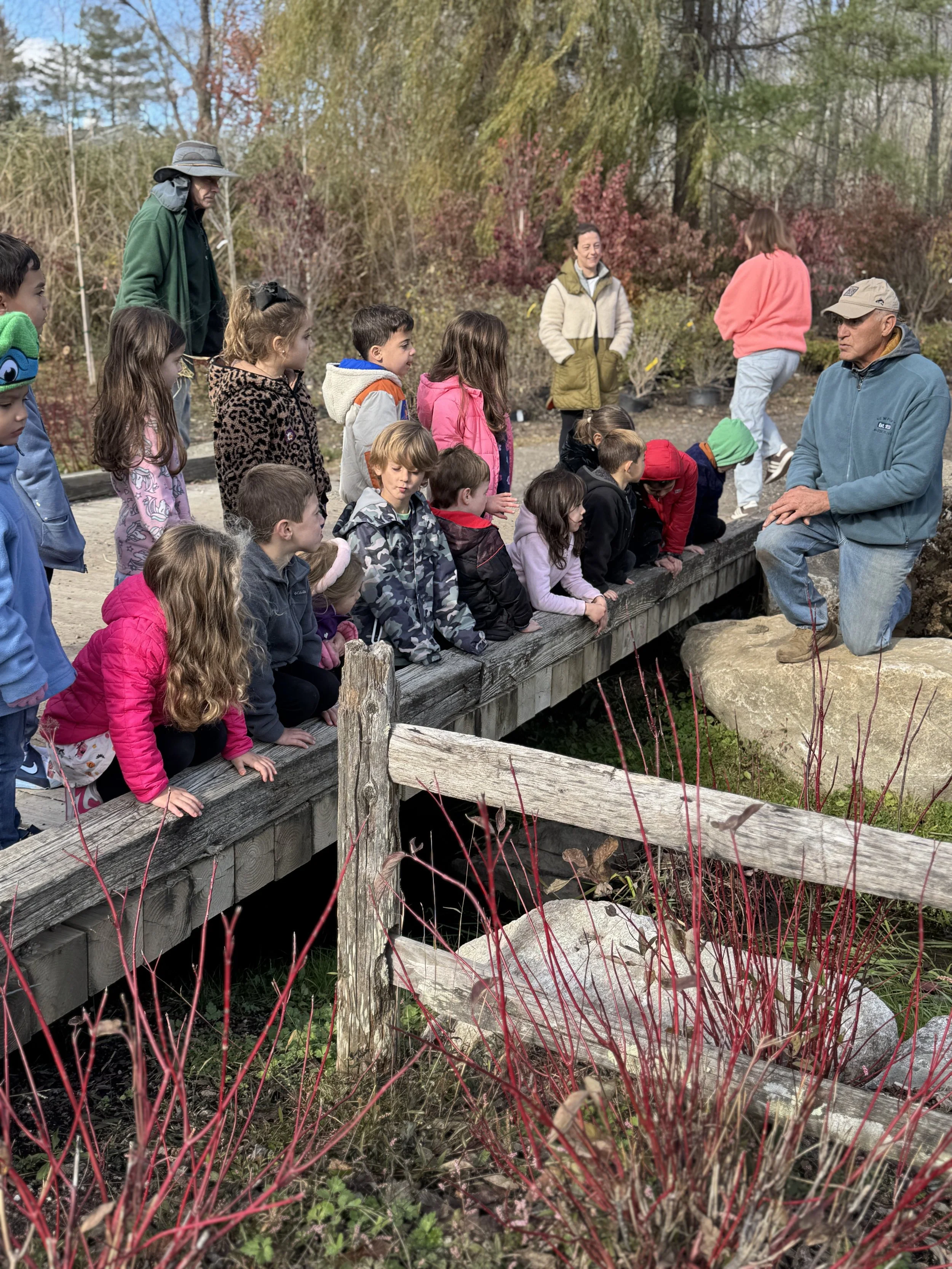 A group of children and a few adults are gathered around a trainer during an outdoor educational visit to a nature park. The children are standing or kneeling on a wooden platform, attentively listening to the man in a tan cap and blue jacket. The scene is set in a landscaped area with plants, bushes, and trees, some with pink blossoms, suggesting spring.