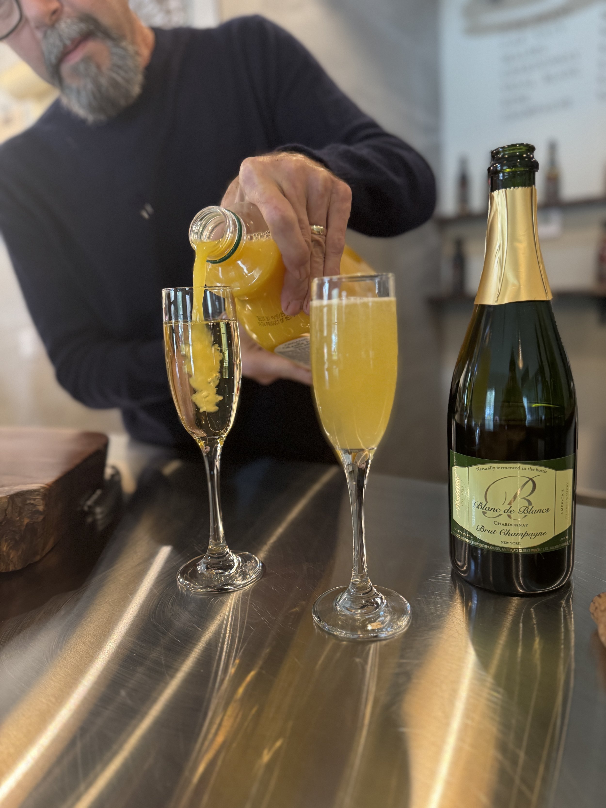 A person pours orange juice into a champagne flute, with a bottle of sparkling wine nearby on a reflective metal surface.