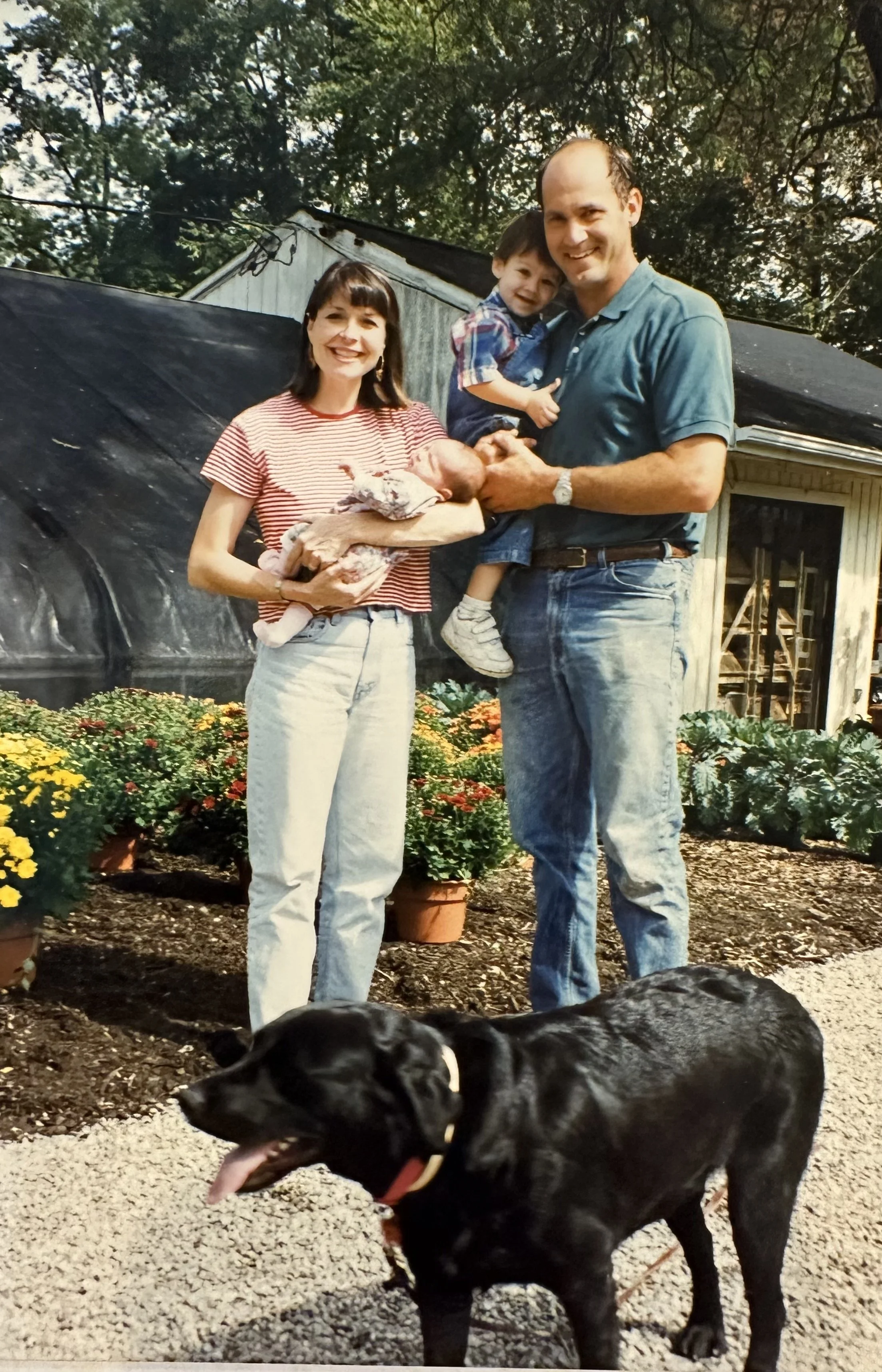 A family of four standing outside in a garden with blooming flowers and trees in the background. The mother is holding a newborn baby, the father is holding a young boy, and a black Labrador dog is in the foreground. The family looks happy and is smiling.